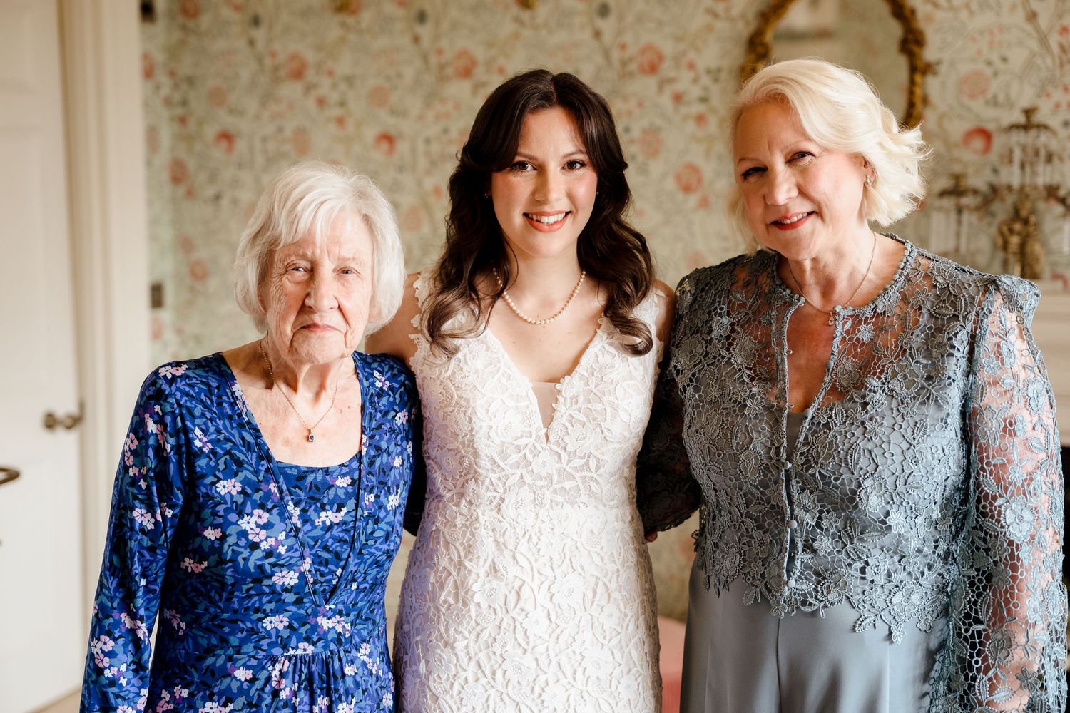Bride in white lace gown poses with two elderly women in blue and gray dresses at a wedding celebration.