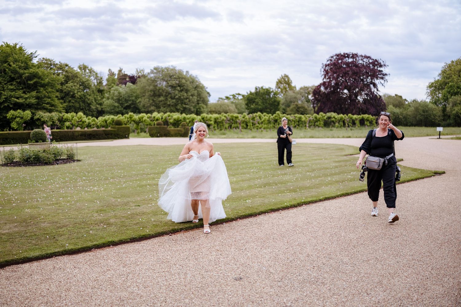 Bride in white dress running across manicured garden lawn with guests following at an elegant outdoor wedding venue.