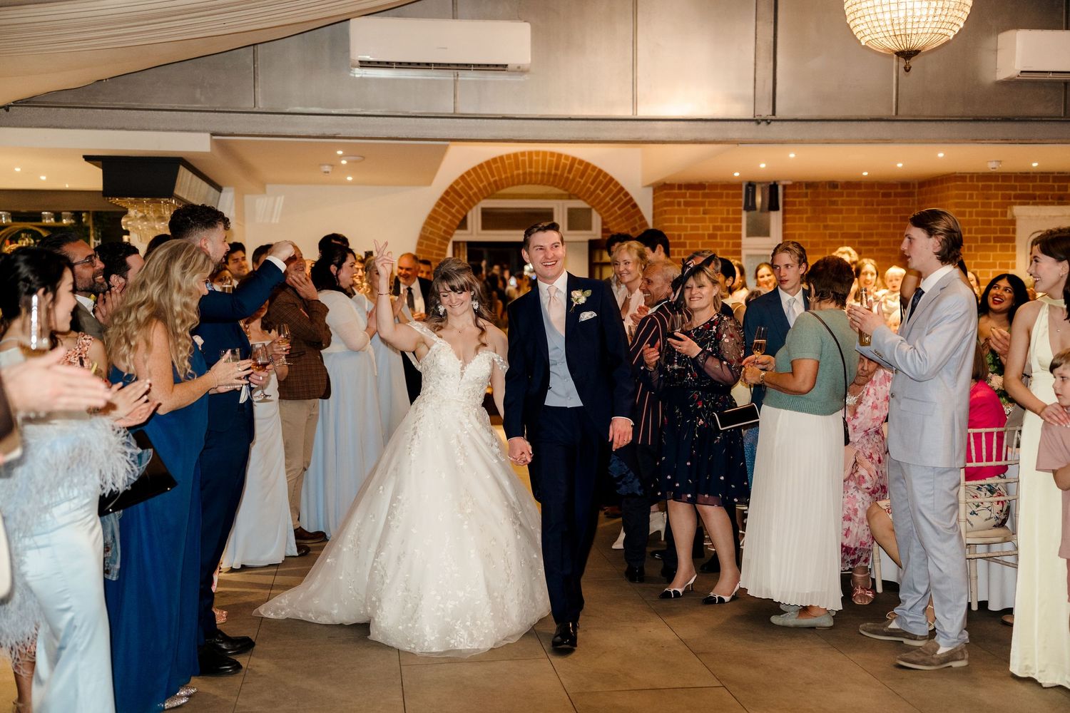 Bride and groom make grand entrance through cheering wedding guests in a elegant brick-walled reception venue.