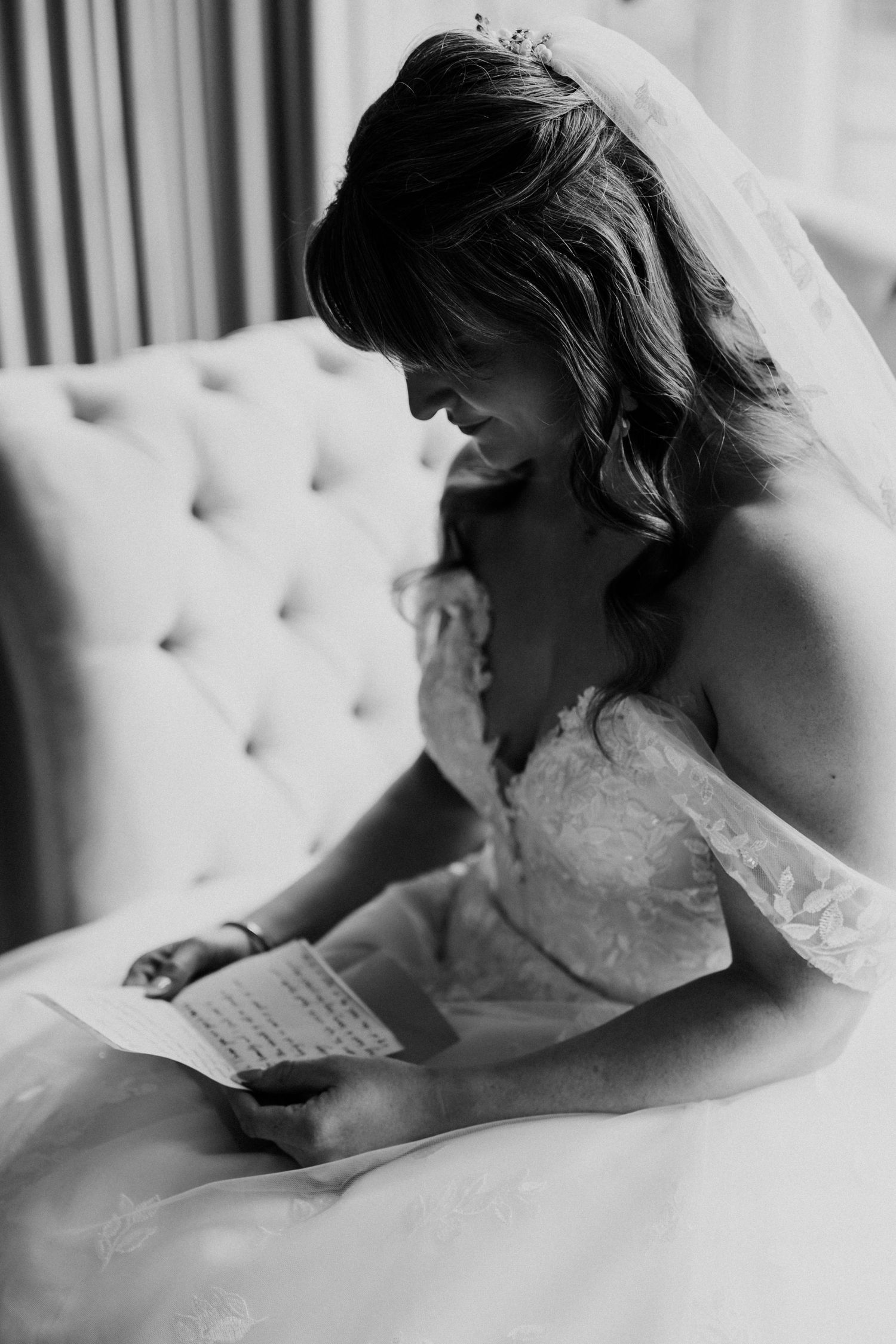 Black and white portrait of bride in lace gown reading a heartfelt letter while seated on tufted sofa.