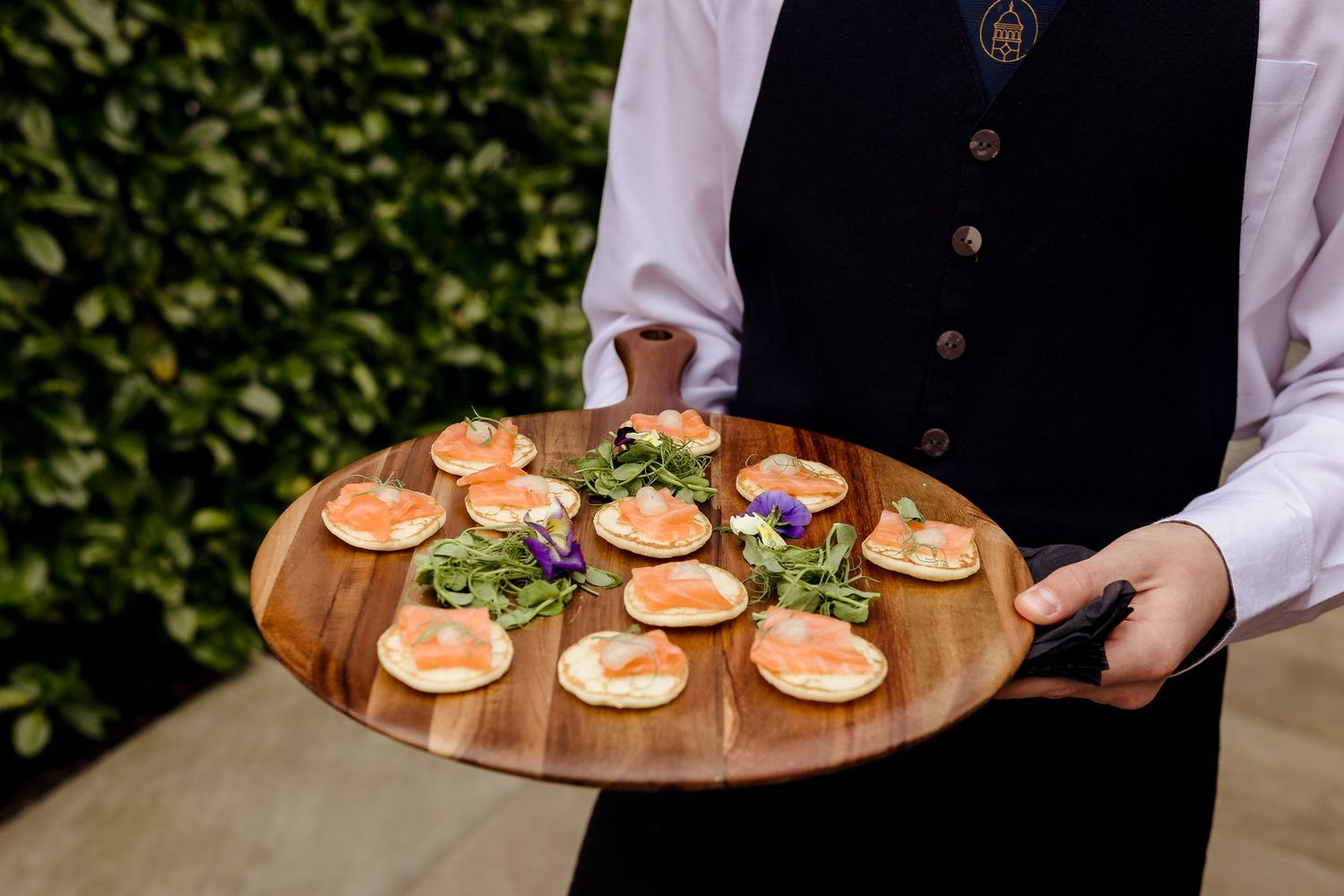 Waiter in black apron serving elegant smoked salmon blini canapes on a round wooden board outdoors.