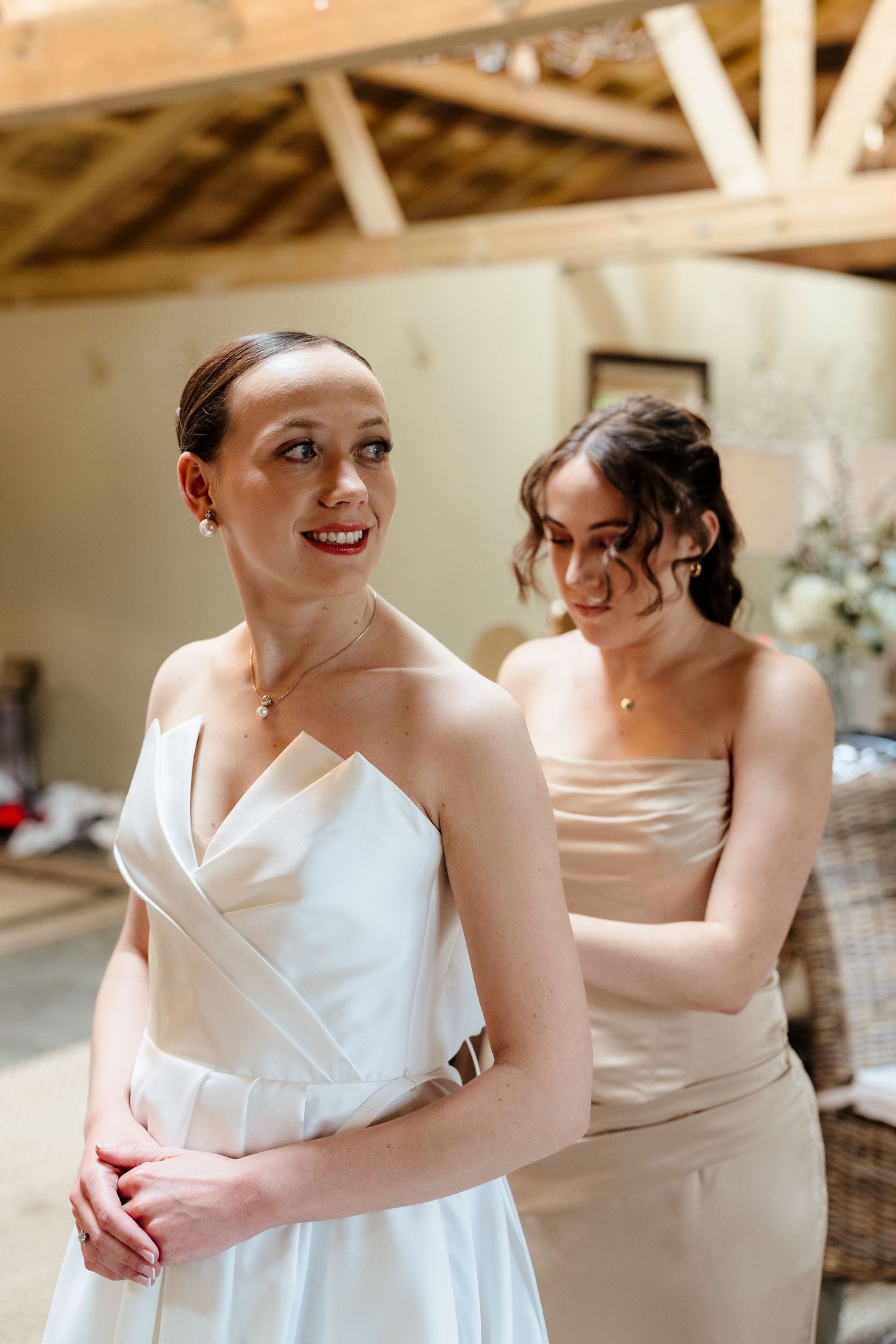Bride in white gown gets help from bridesmaid in champagne dress inside rustic wooden venue.