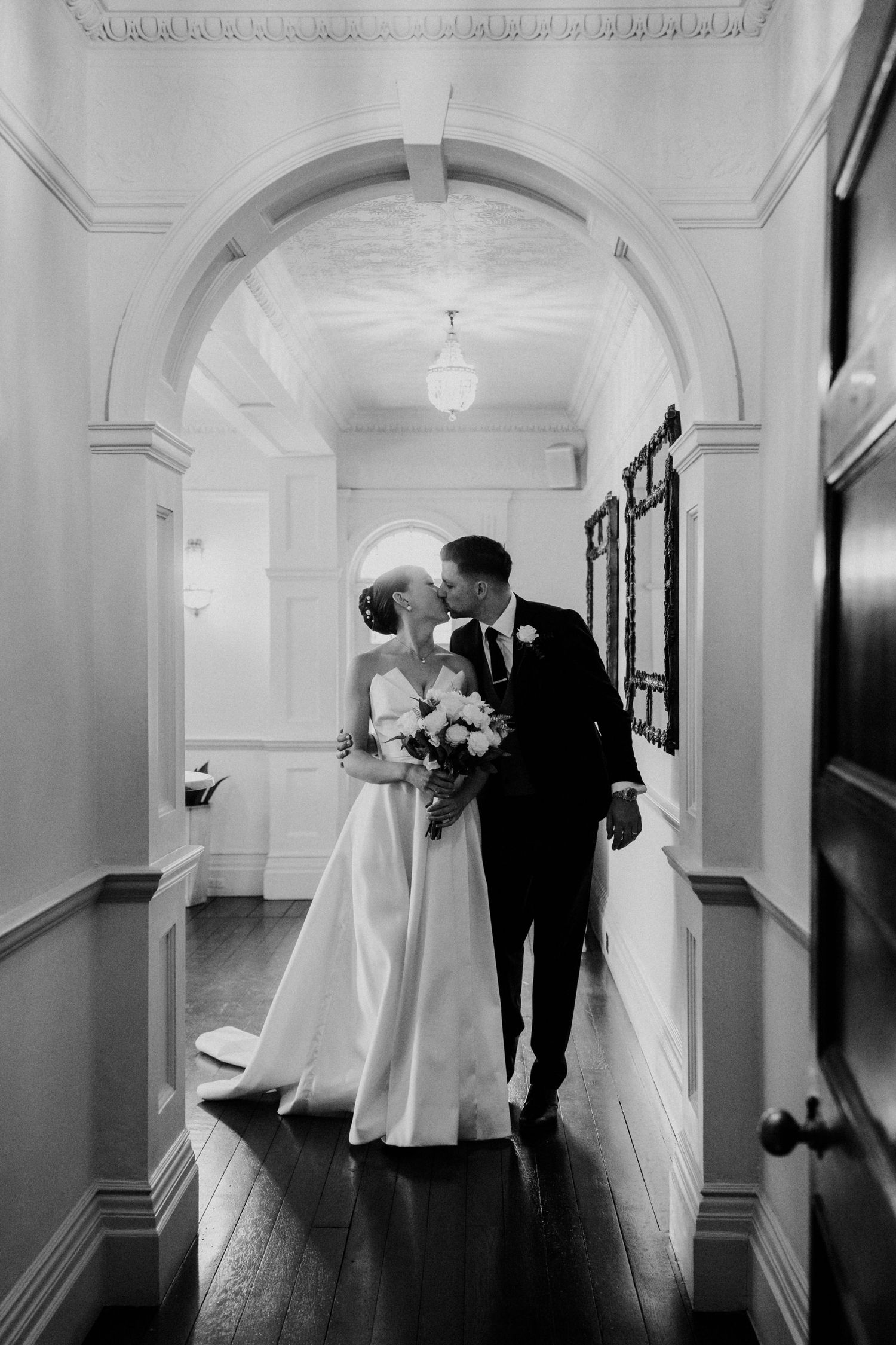 Bride and groom share a kiss in elegant hallway in timeless black and white wedding photo.