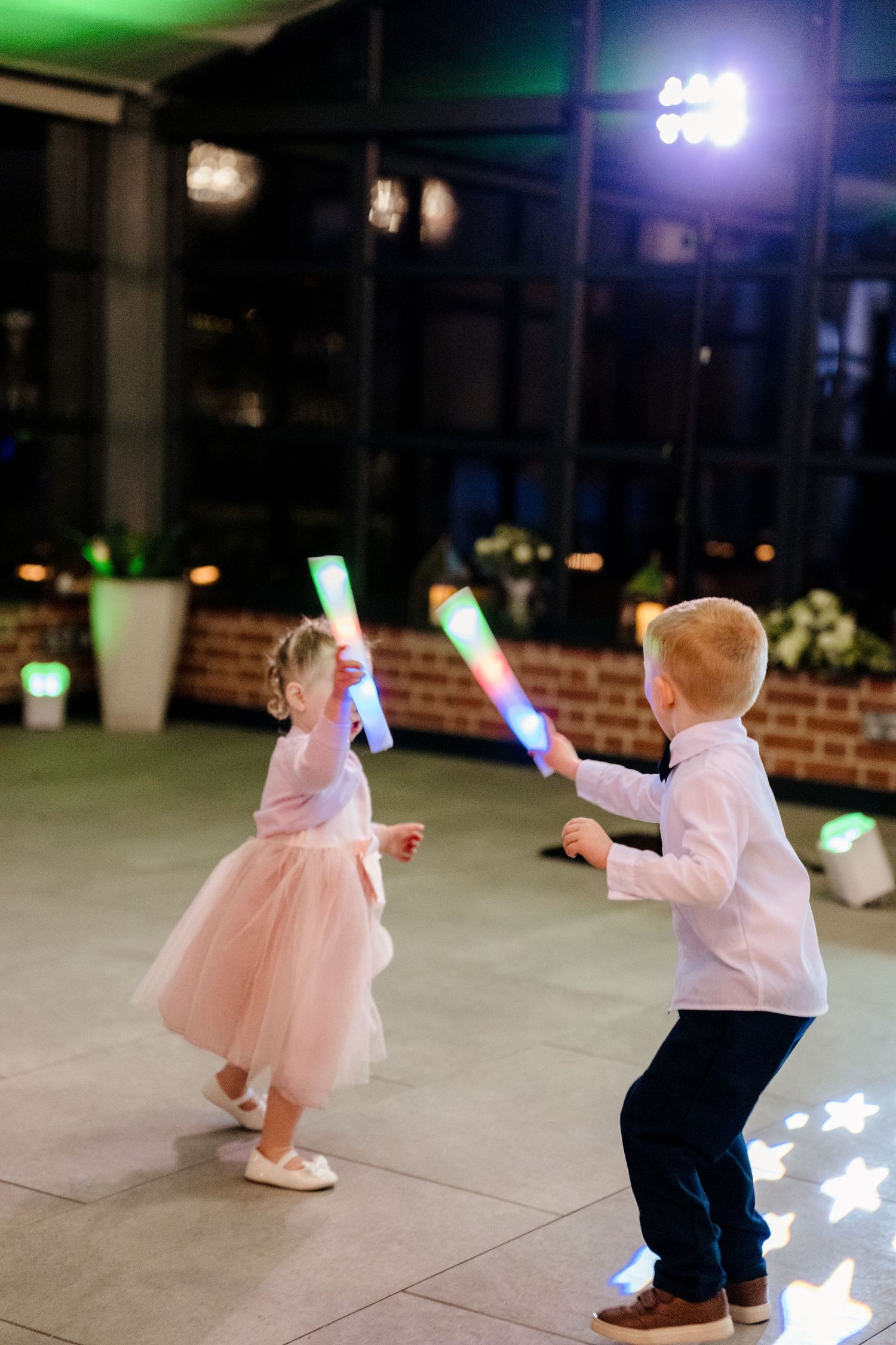 Two young children playing with colorful LED foam sticks on a wedding reception dance floor at night.