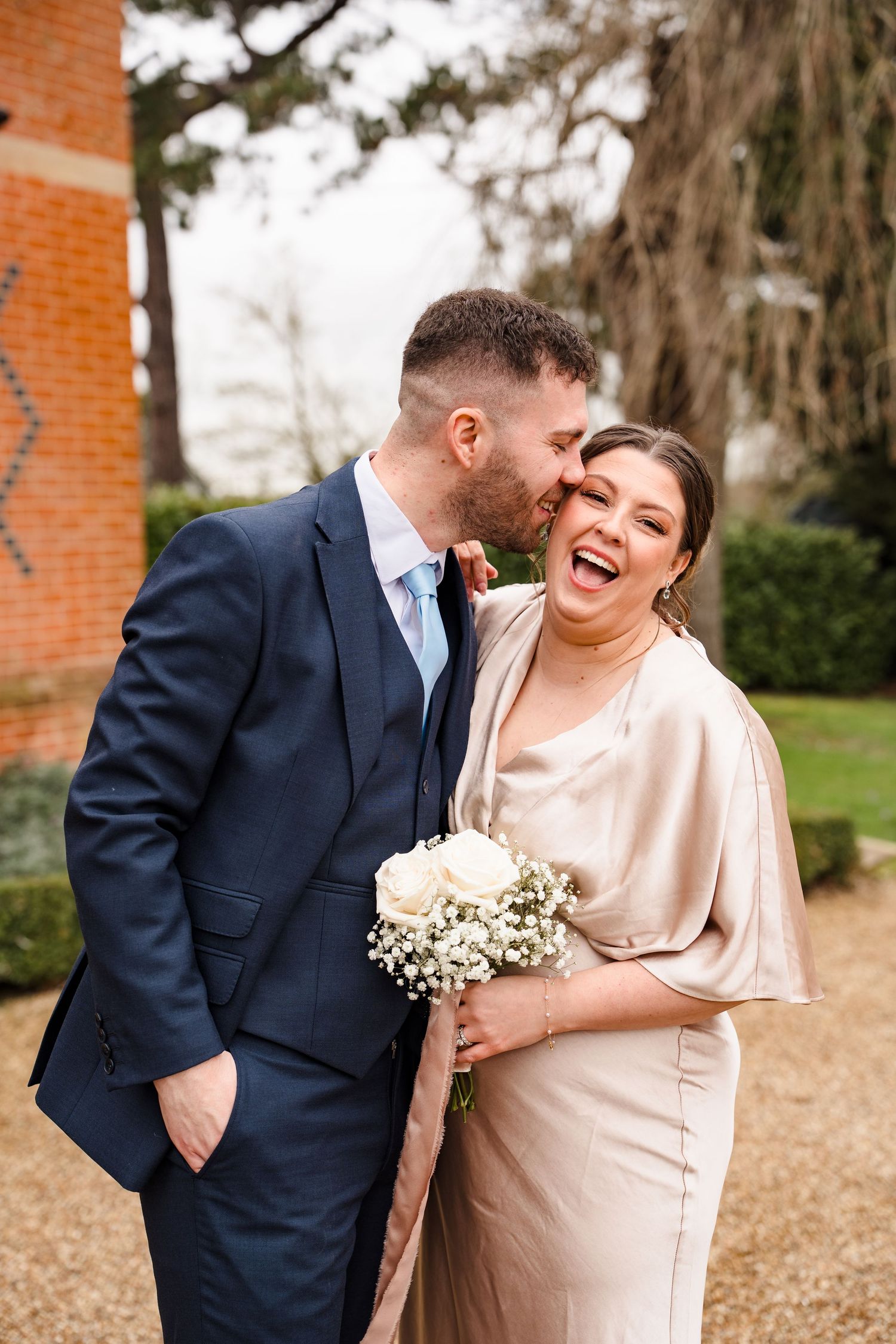 Groom in navy suit playfully whispering in laughing bride's ear outdoors at a brick manor wedding venue.