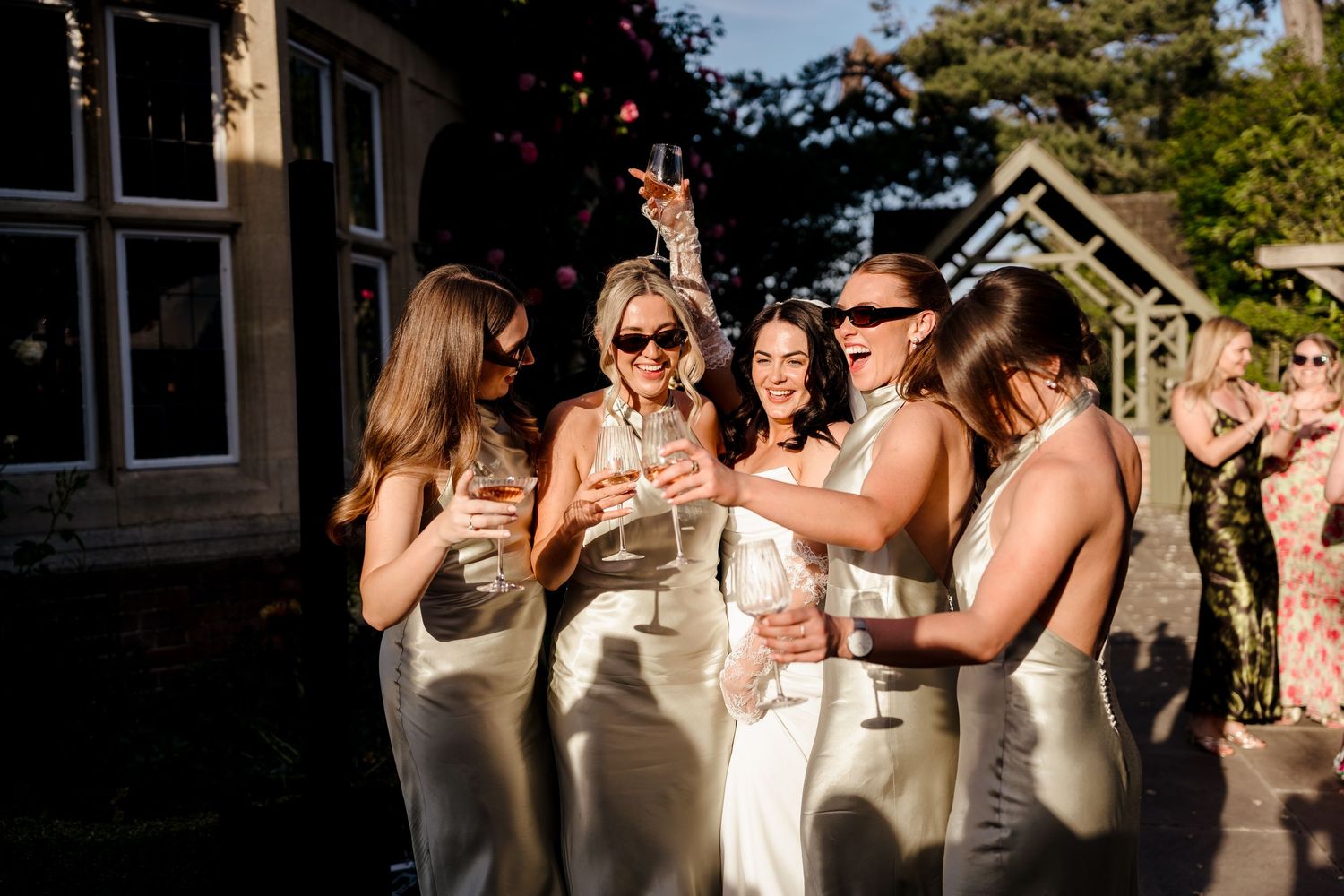 Four bridesmaids in champagne satin dresses celebrate outdoors with champagne glasses in golden sunlight.