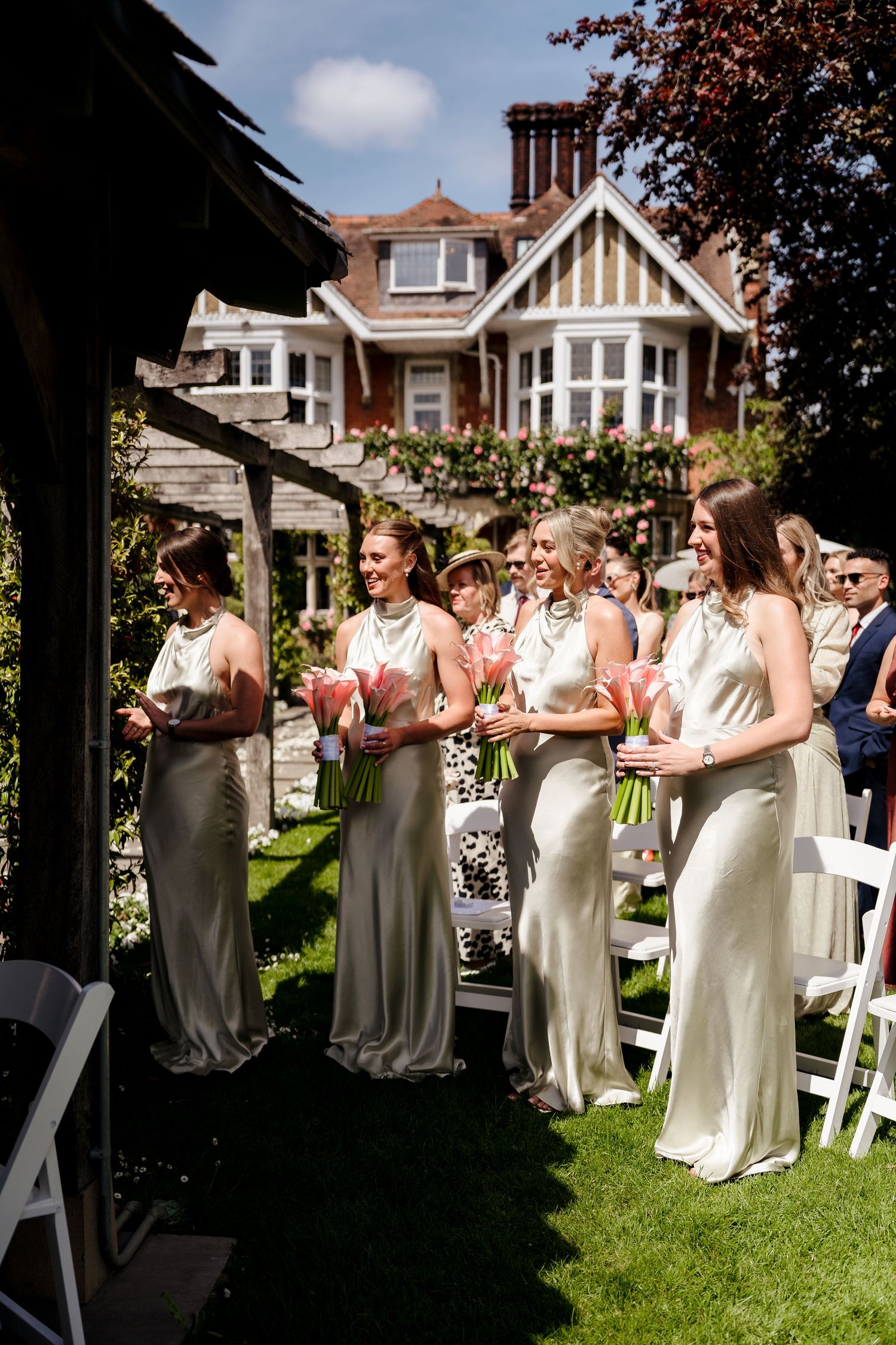 Bridesmaids in elegant silver satin gowns holding pink tulip bouquets stand at an outdoor wedding ceremony beside a grand manor.