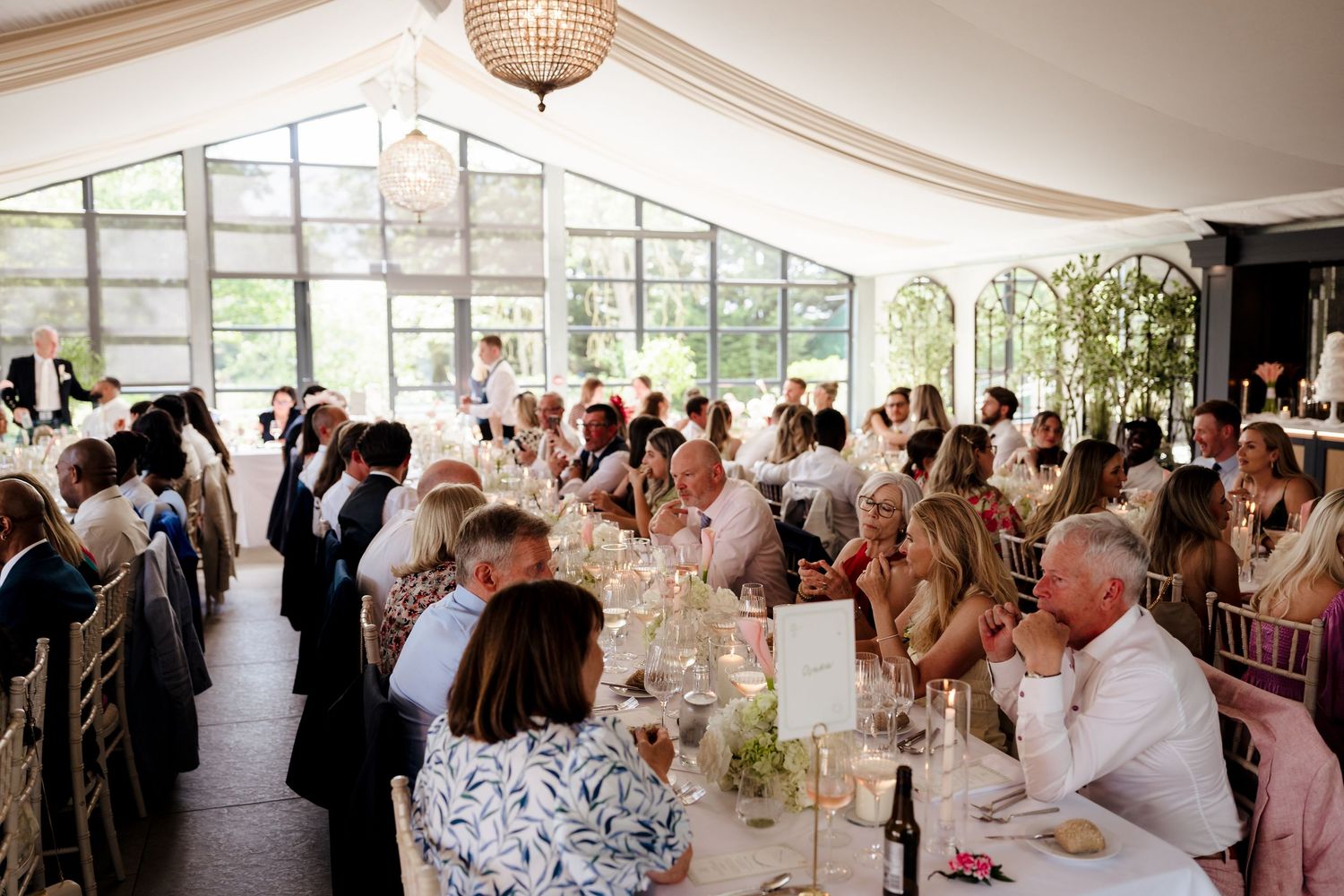 Elegant wedding reception in glass marquee with guests seated at long tables decorated with florals.