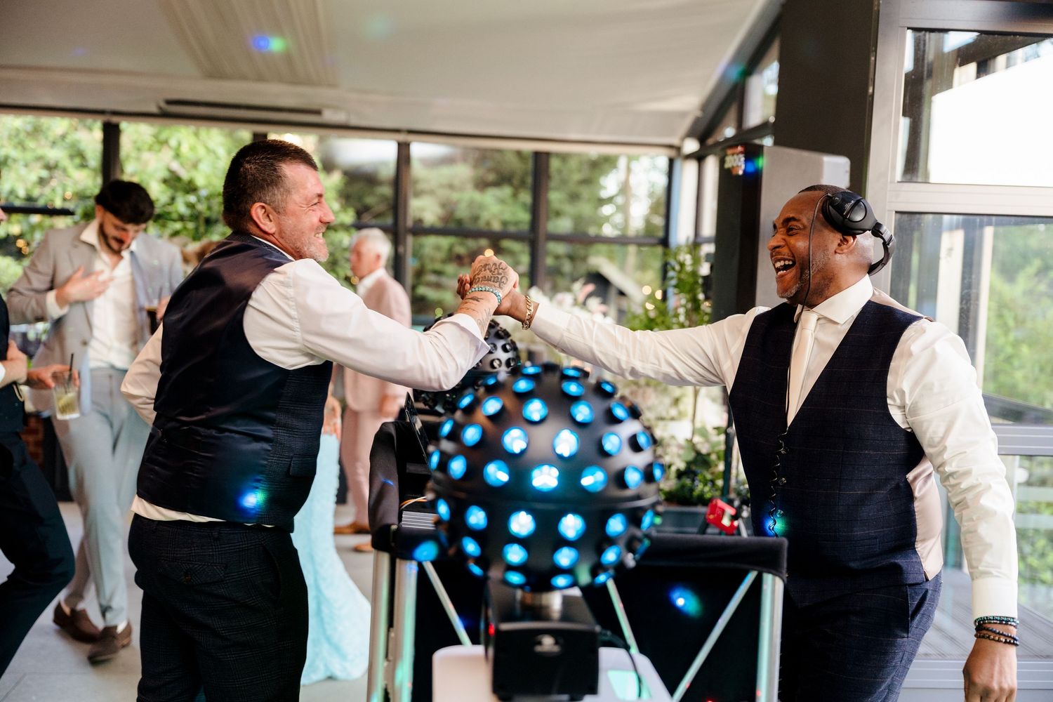 DJ and guest share an enthusiastic fist bump beside a disco ball at a lively wedding reception.