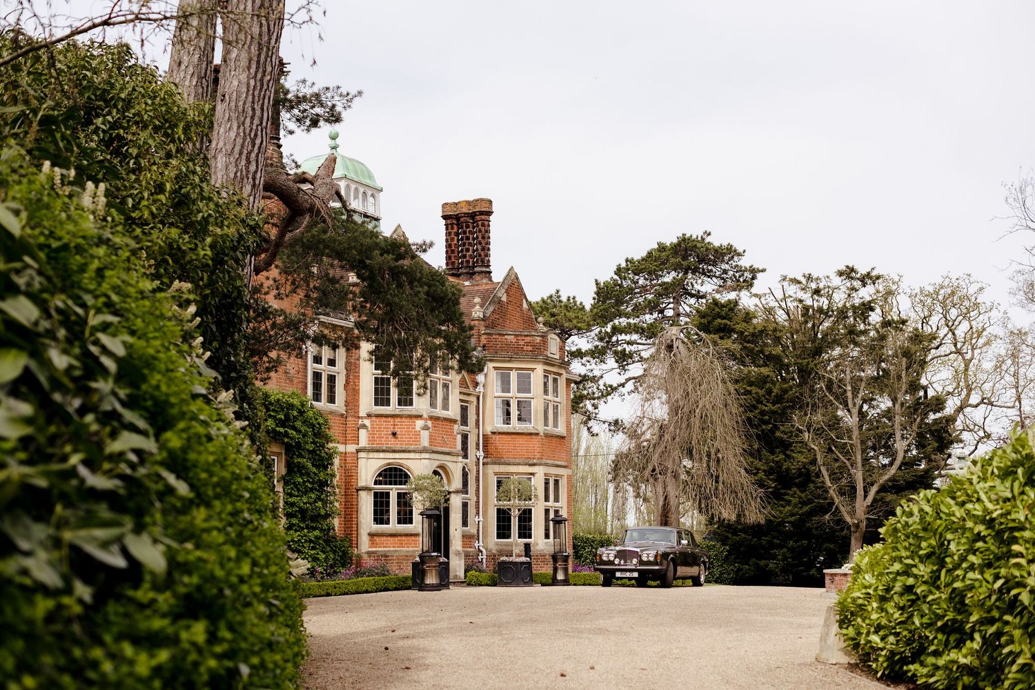 A grand Victorian red brick manor house surrounded by lush green hedges and tall trees on a gravel driveway.