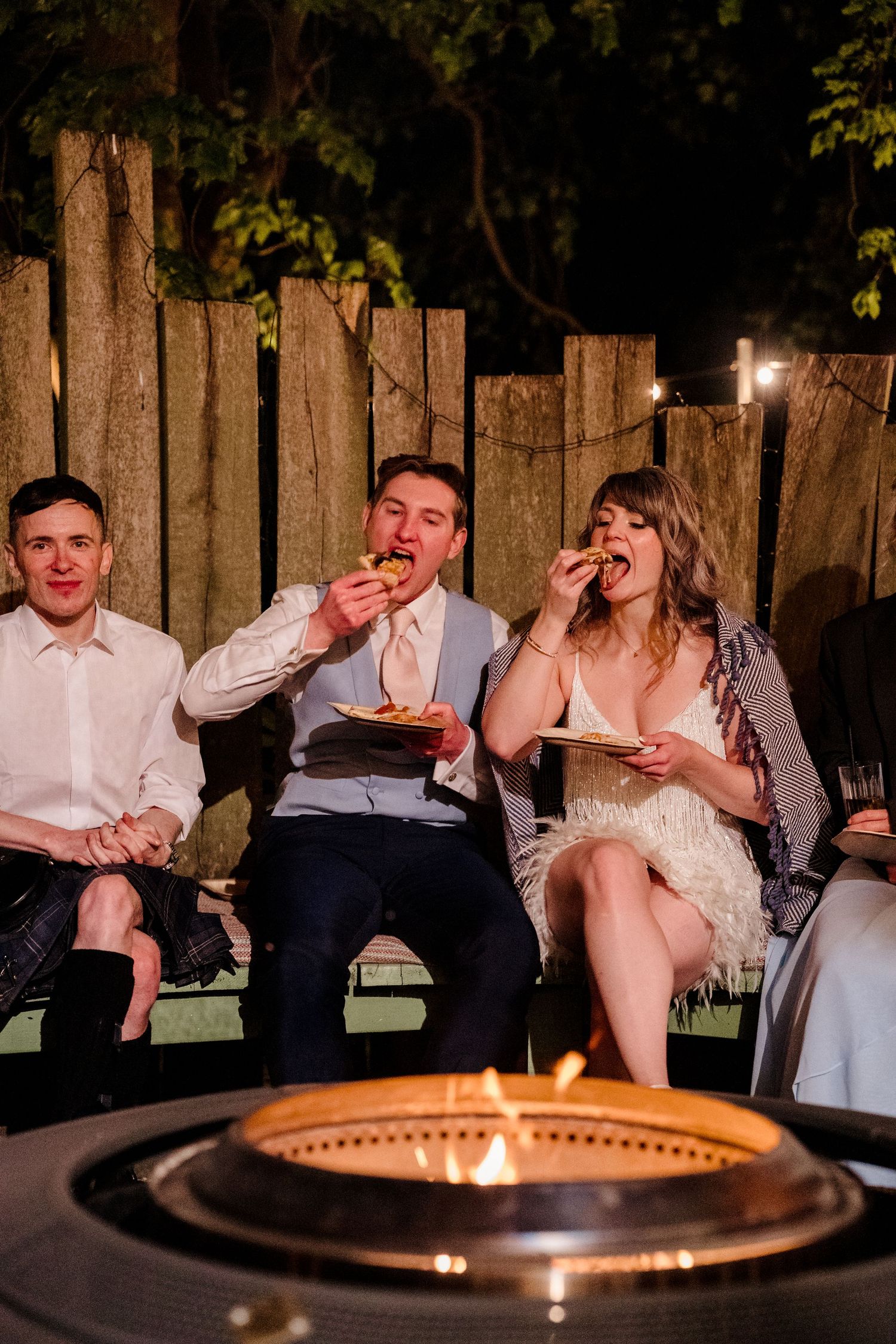Bride and groom eating late night snacks with a guest beside a fire pit at an outdoor wedding reception.