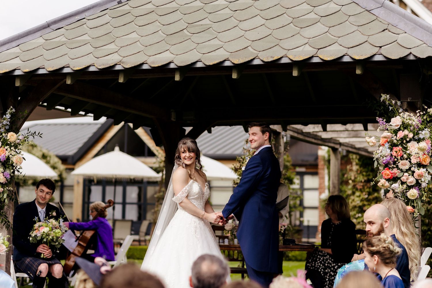 Bride and groom smile during outdoor wedding ceremony under floral arch gazebo.
