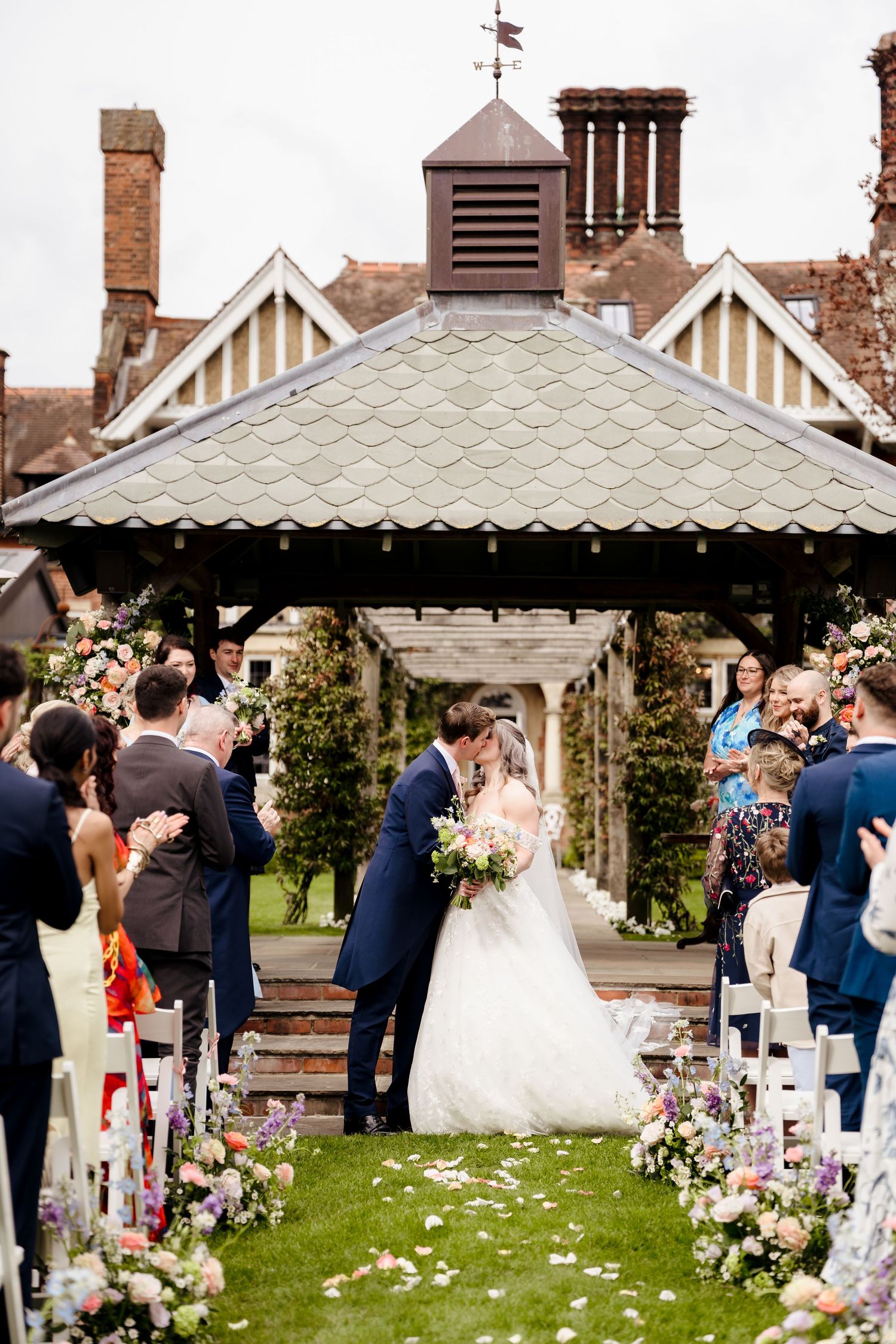 Bride and groom sharing first kiss at outdoor ceremony aisle lined with colorful floral arrangements.