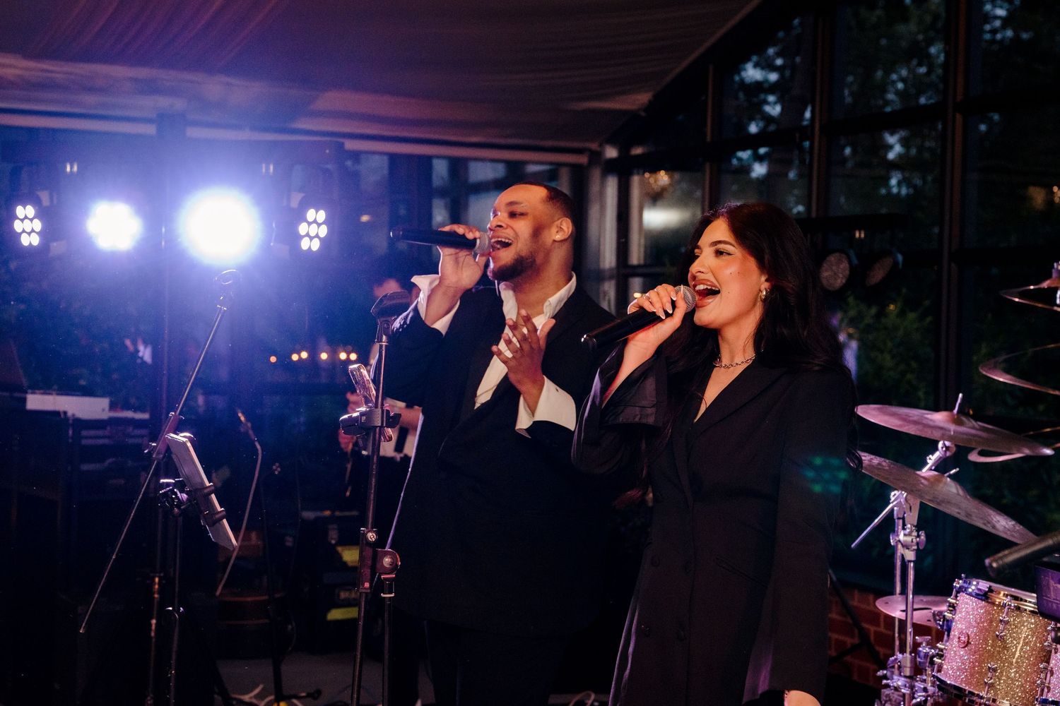 Male and female wedding band singers perform on stage with microphones at evening wedding reception.