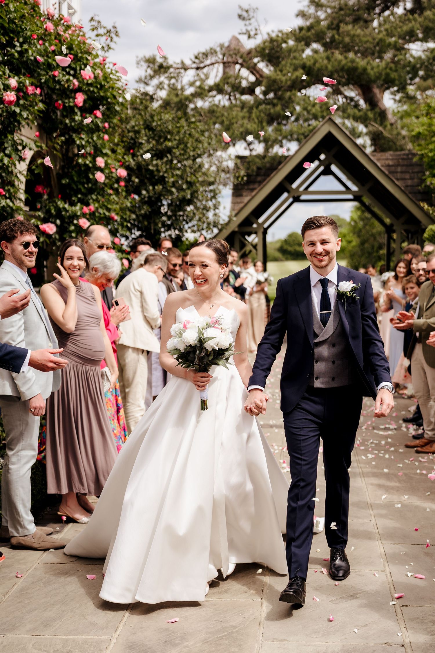 Joyful bride and groom walking through confetti shower lined by cheering guests after wedding ceremony.