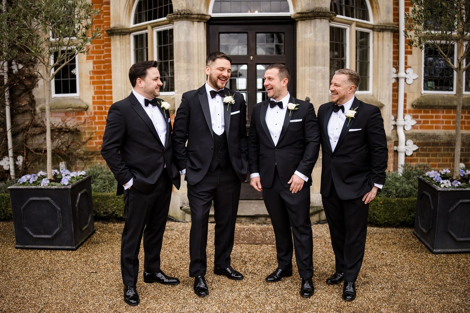 Four groomsmen in black tuxedos with bow ties laughing together outside an elegant brick venue.
