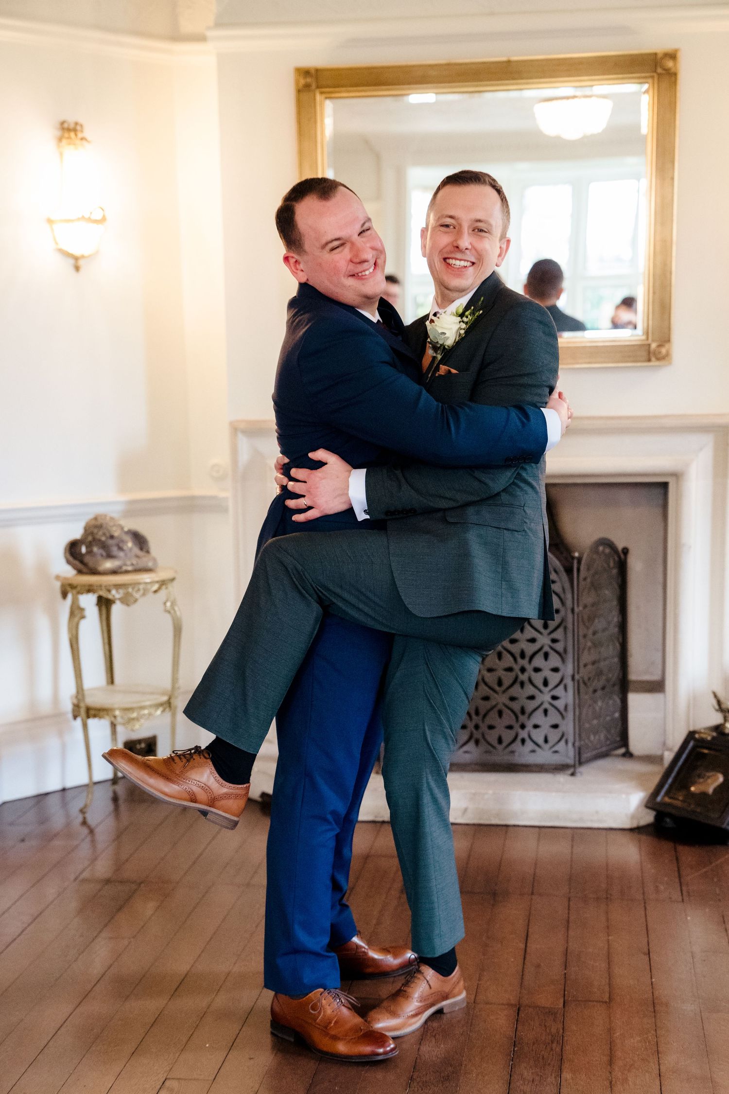 Two grooms share a joyful embrace in an elegant room with fireplace at their wedding celebration.