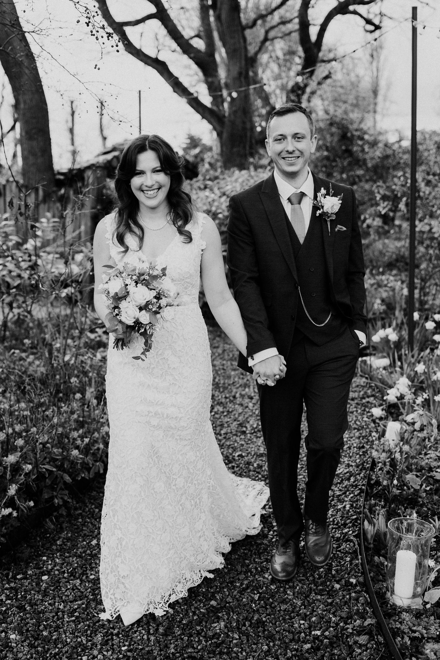 Black and white photo of smiling bride and groom holding hands walking through garden aisle.