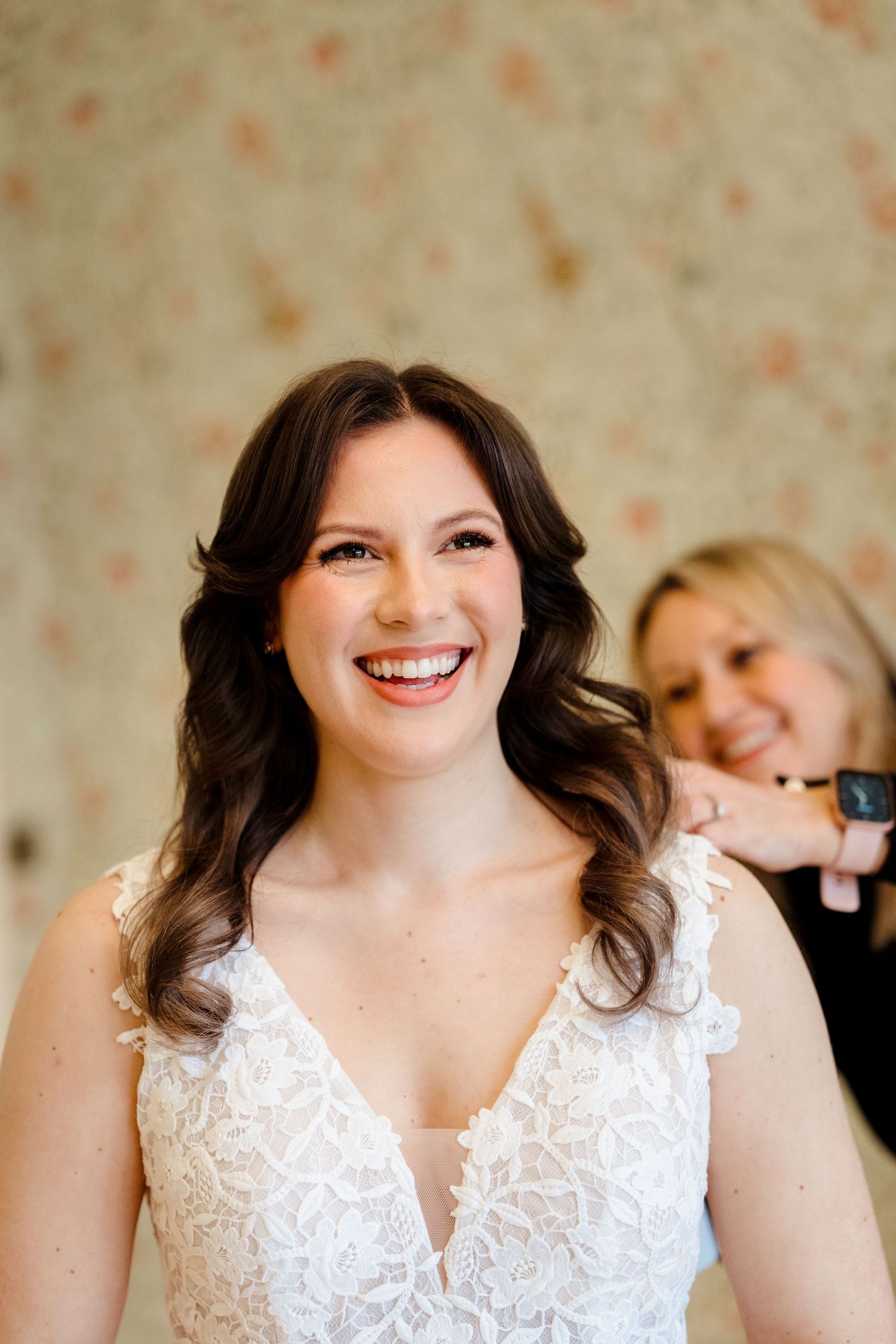 Radiant bride in lace wedding dress smiling joyfully while bridesmaid helps with final preparations.