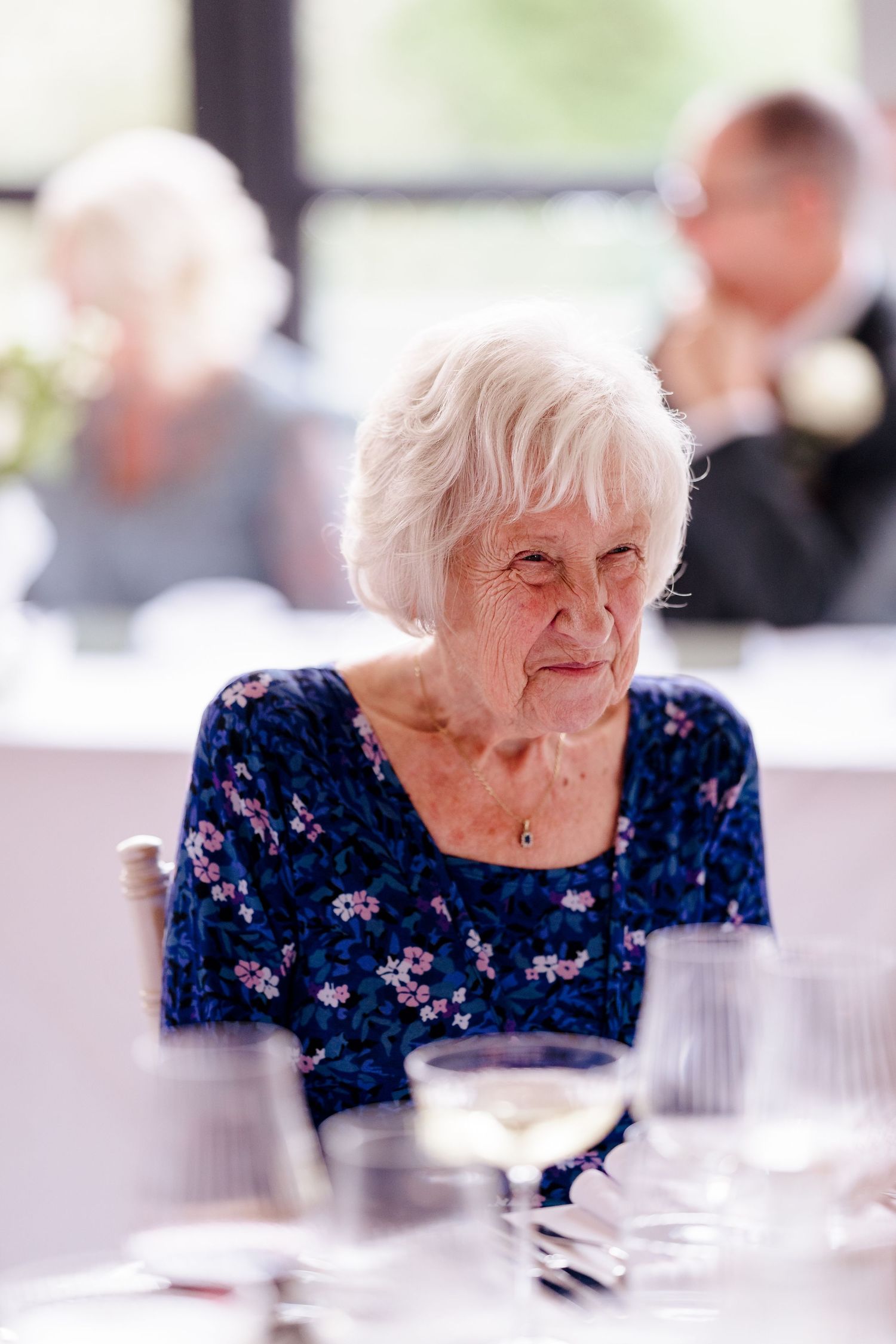 Elegant elderly woman in blue floral dress seated at wedding reception table with wine glasses.