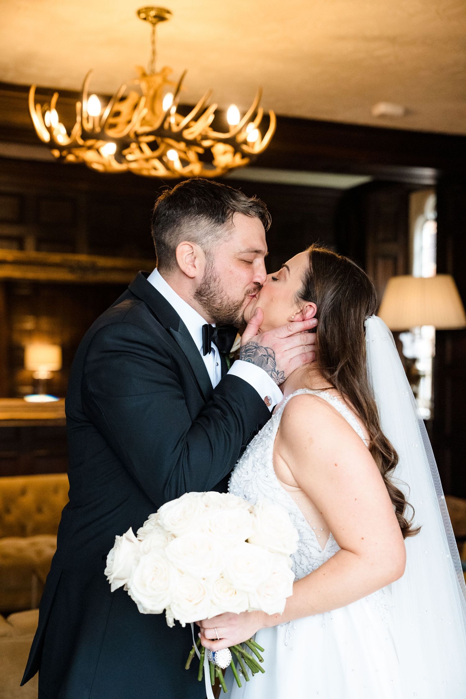 Newlyweds share a kiss as groom holds white rose bouquet beneath an antler chandelier.