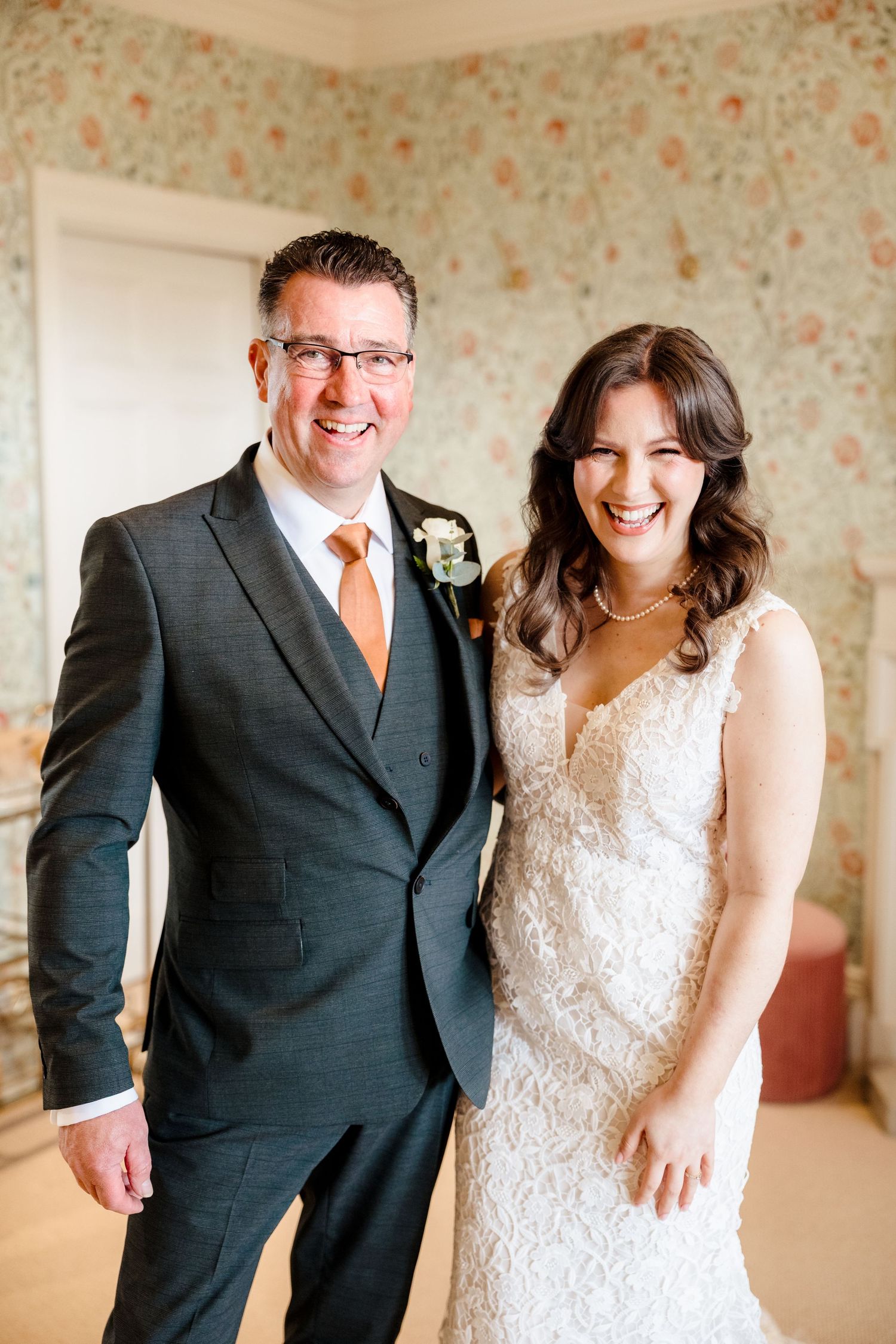 Smiling bride in lace gown poses with older man in grey suit and orange tie.