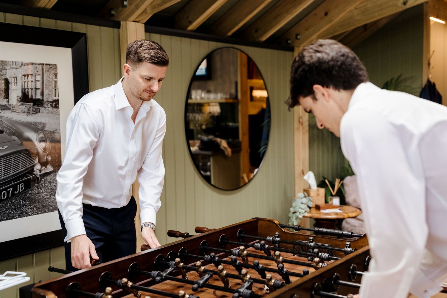 Two men in white shirts playing foosball in a stylish game room with green walls and a round mirror.