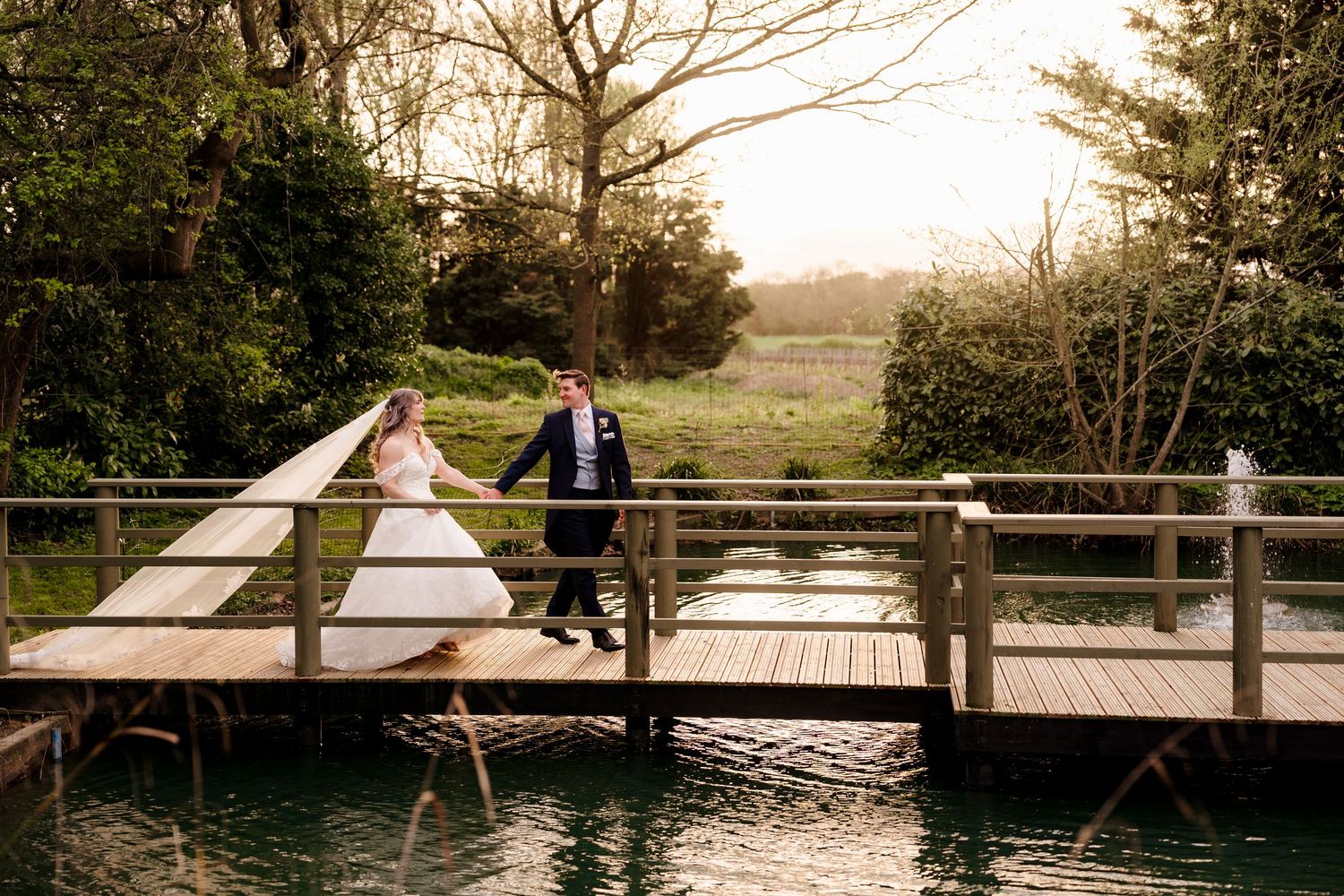 Bride and groom walk hand-in-hand across a wooden bridge at golden hour by a lake.