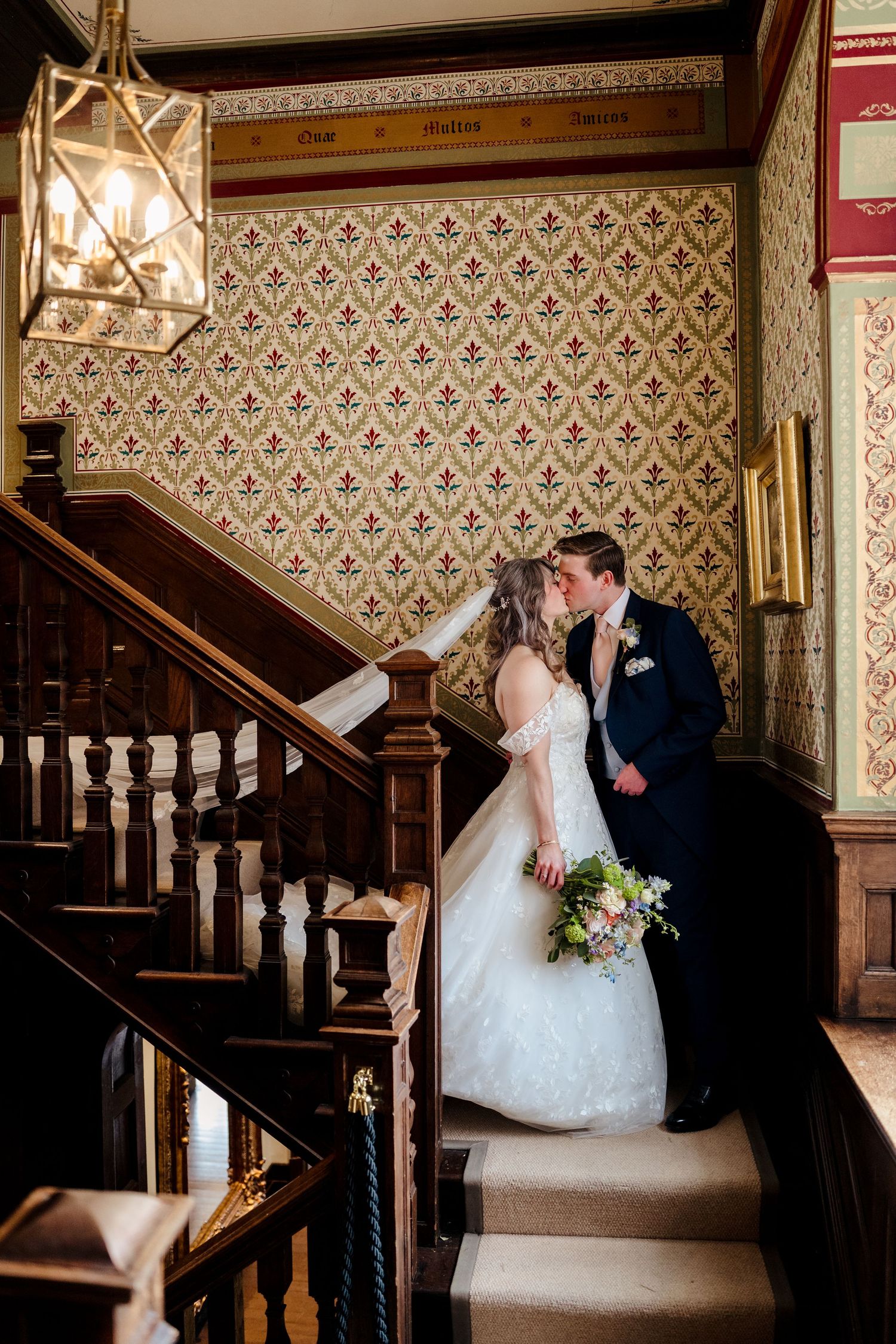 Bride and groom sharing a kiss on ornate wooden staircase with decorative wallpaper backdrop.