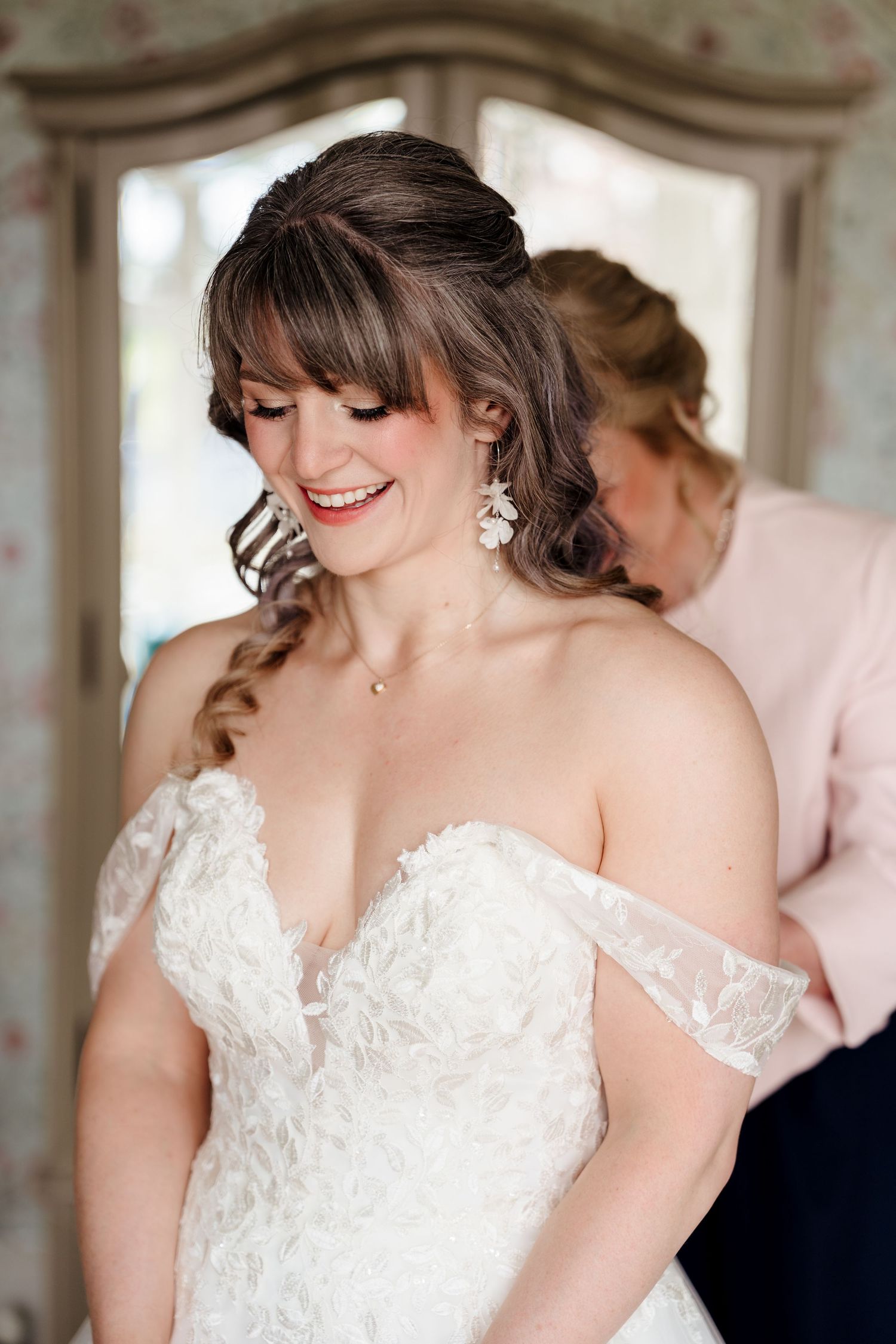 Smiling bride in off-shoulder lace wedding gown with floral earrings being helped to dress by bridesmaid.