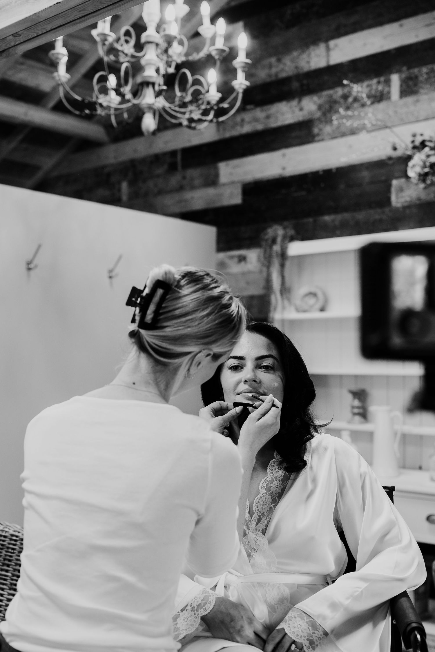 Makeup artist applying lipstick to a bride in a silk robe, black and white photo with chandelier backdrop.