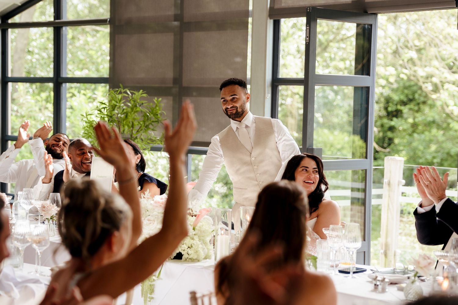 Groom in white vest stands smiling as wedding guests raise hands and applaud in bright reception hall.
