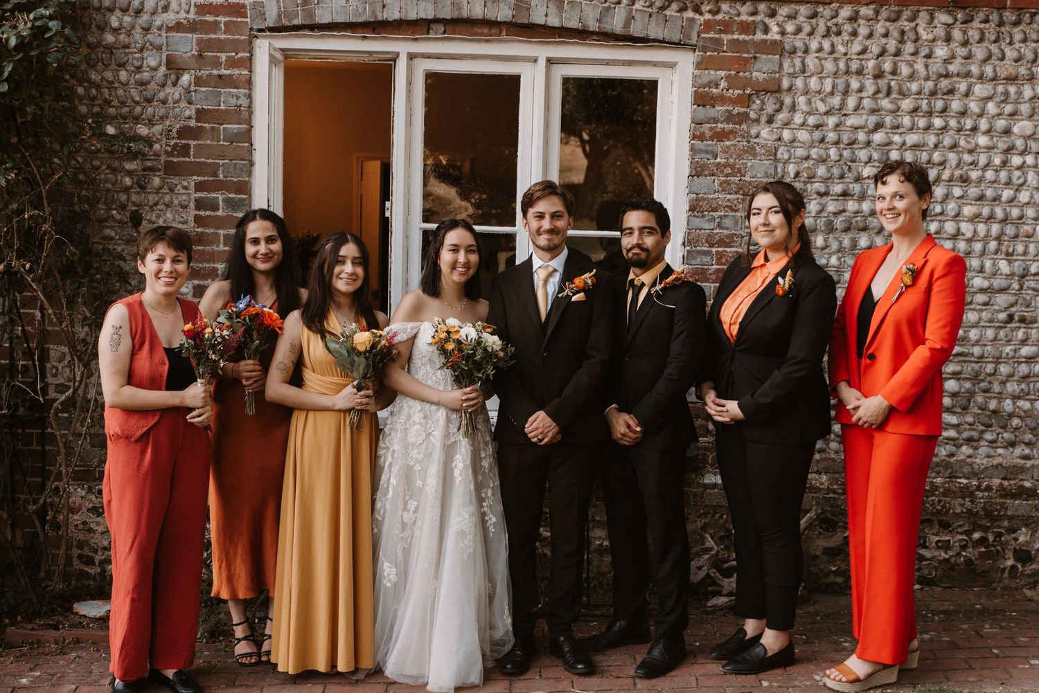 Wedding party poses in autumn tones outside rustic brick venue with bride and groom centered.