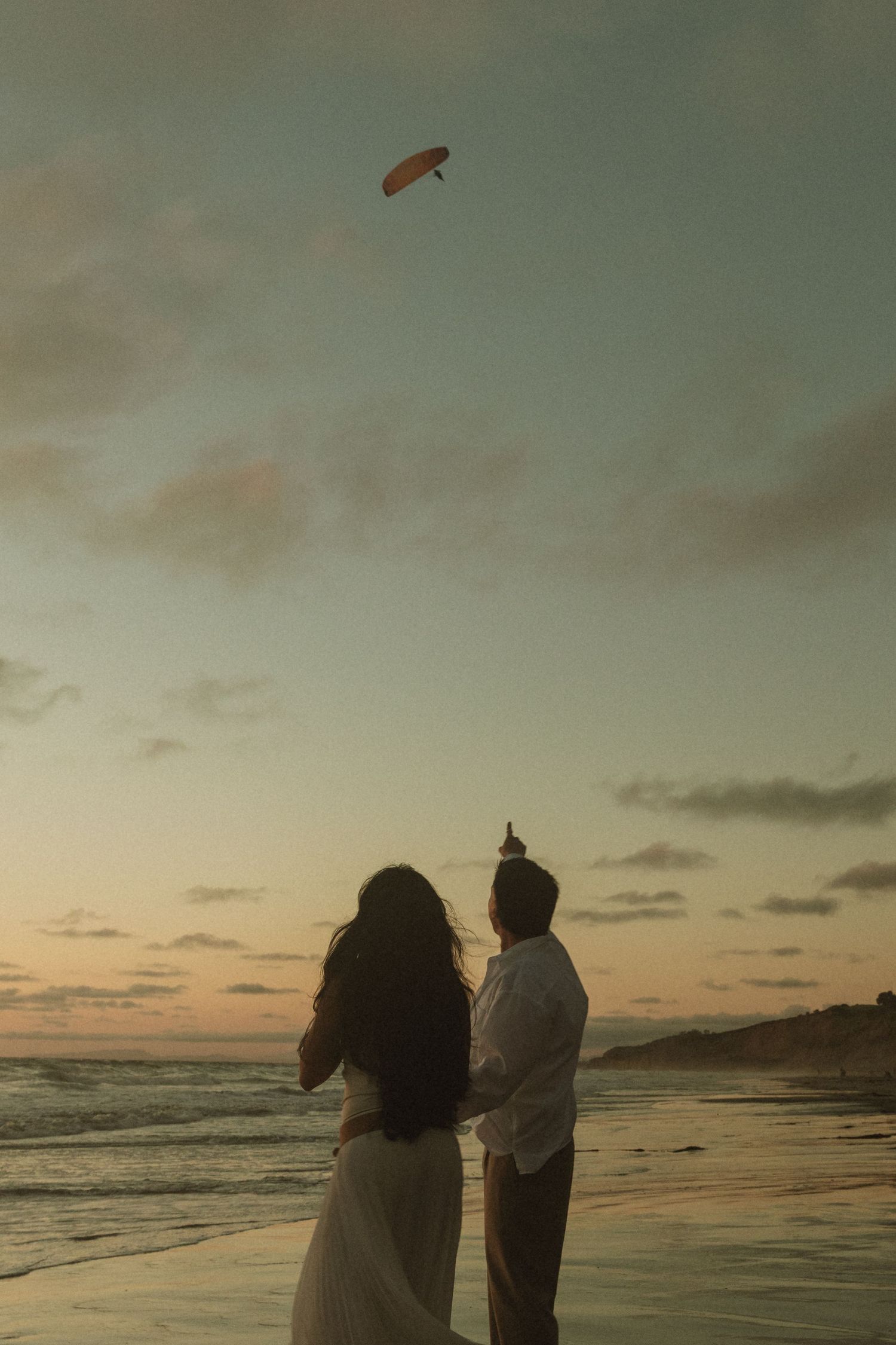 Couple on beach at sunset watching a kite fly overhead, romantic seaside scene with golden sky.