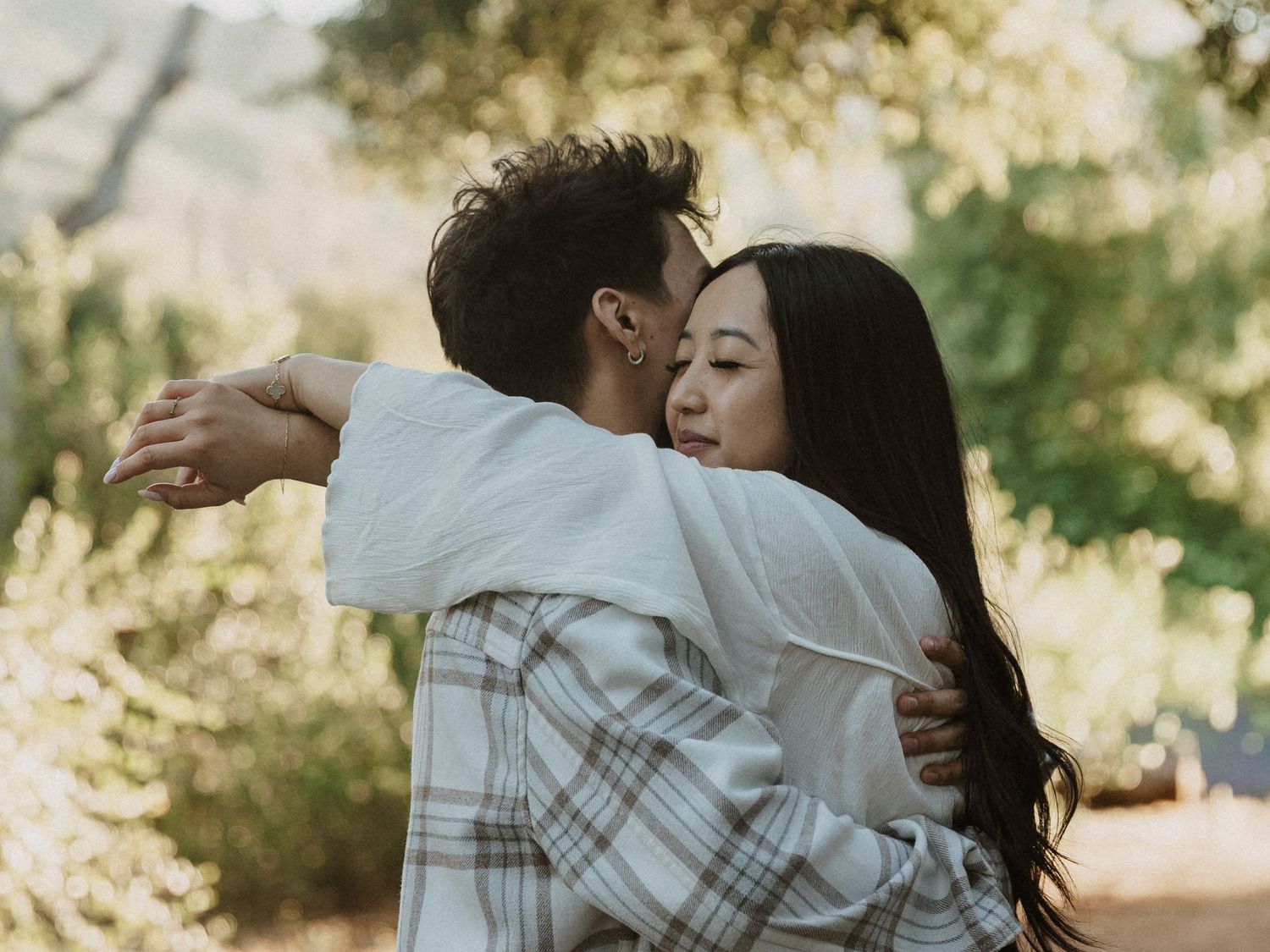 A couple shares a tender embrace outdoors, surrounded by lush green trees in a romantic natural setting.