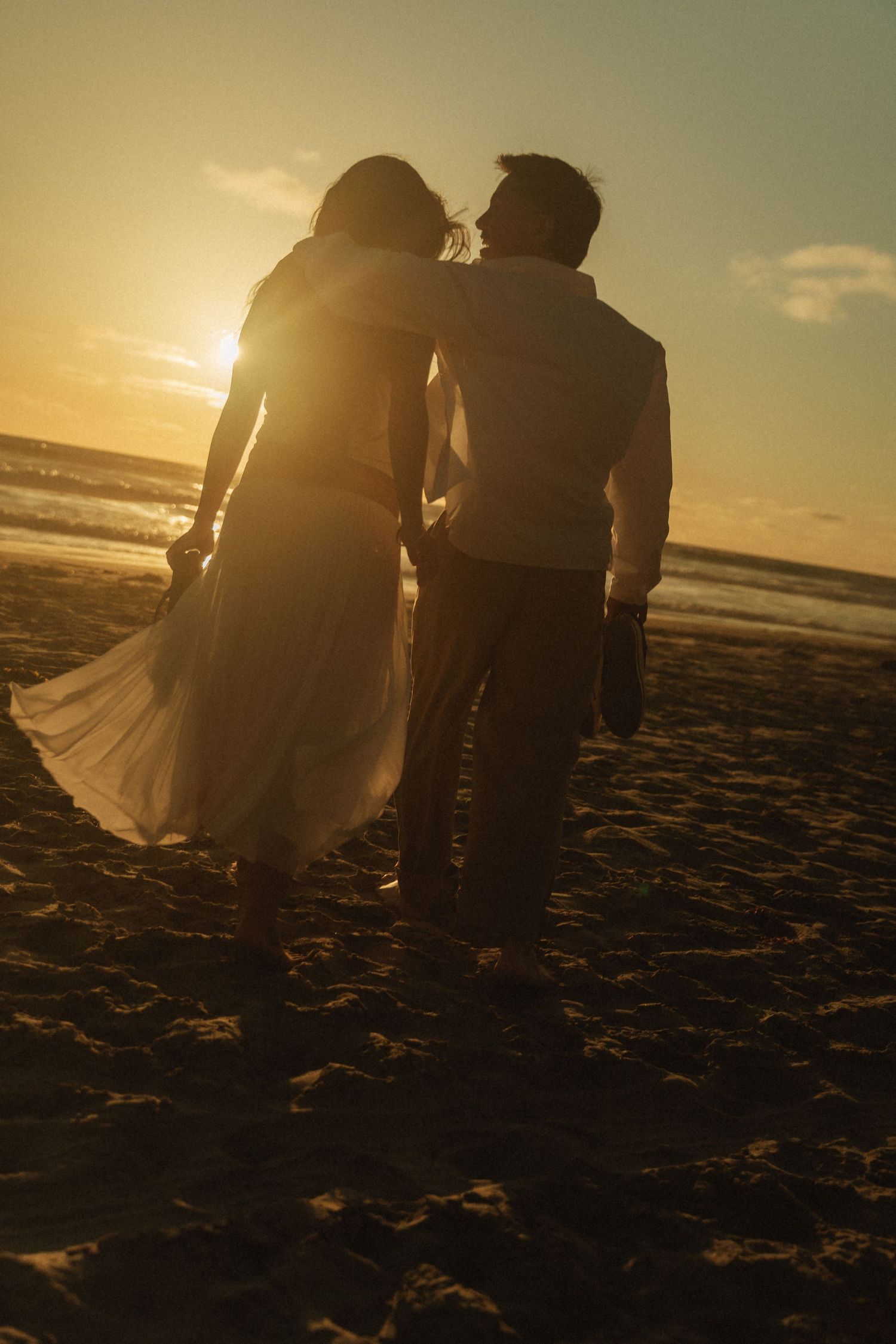 Silhouette of a couple sharing a romantic moment on a sandy beach at golden sunset, woman in flowing dress.