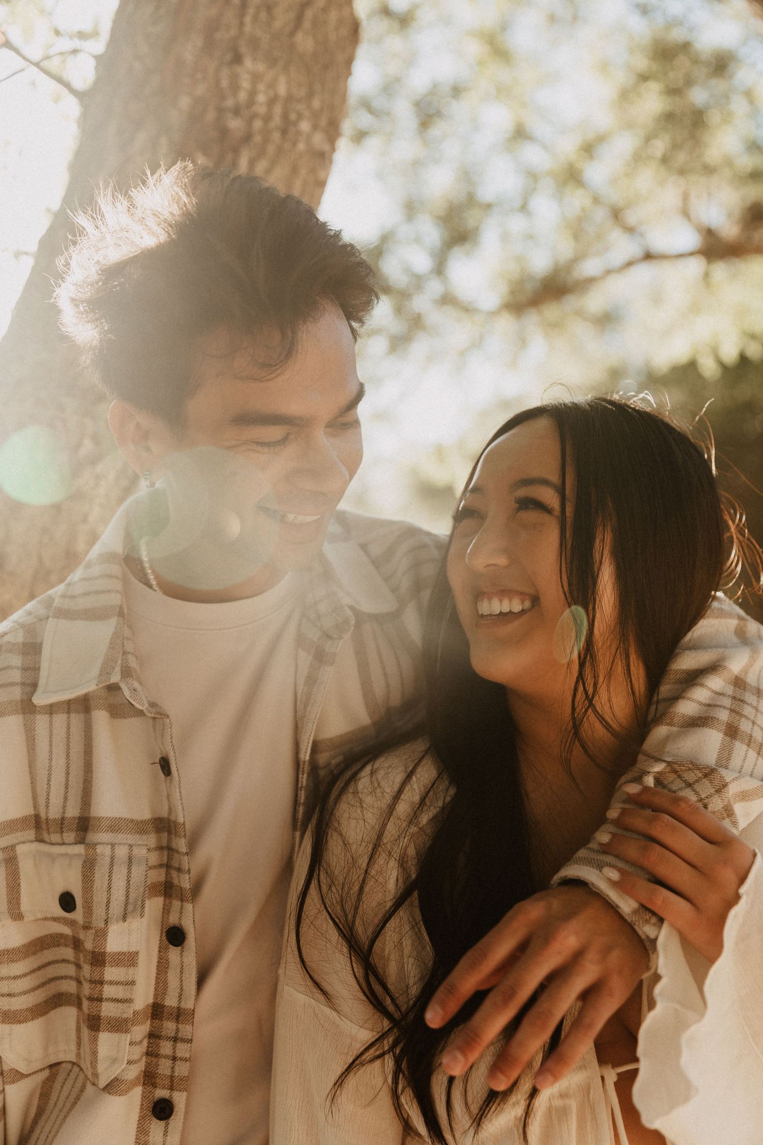 A smiling couple embraces outdoors in warm sunlight, the man in a plaid shirt with his arm around a laughing dark-haired woman.