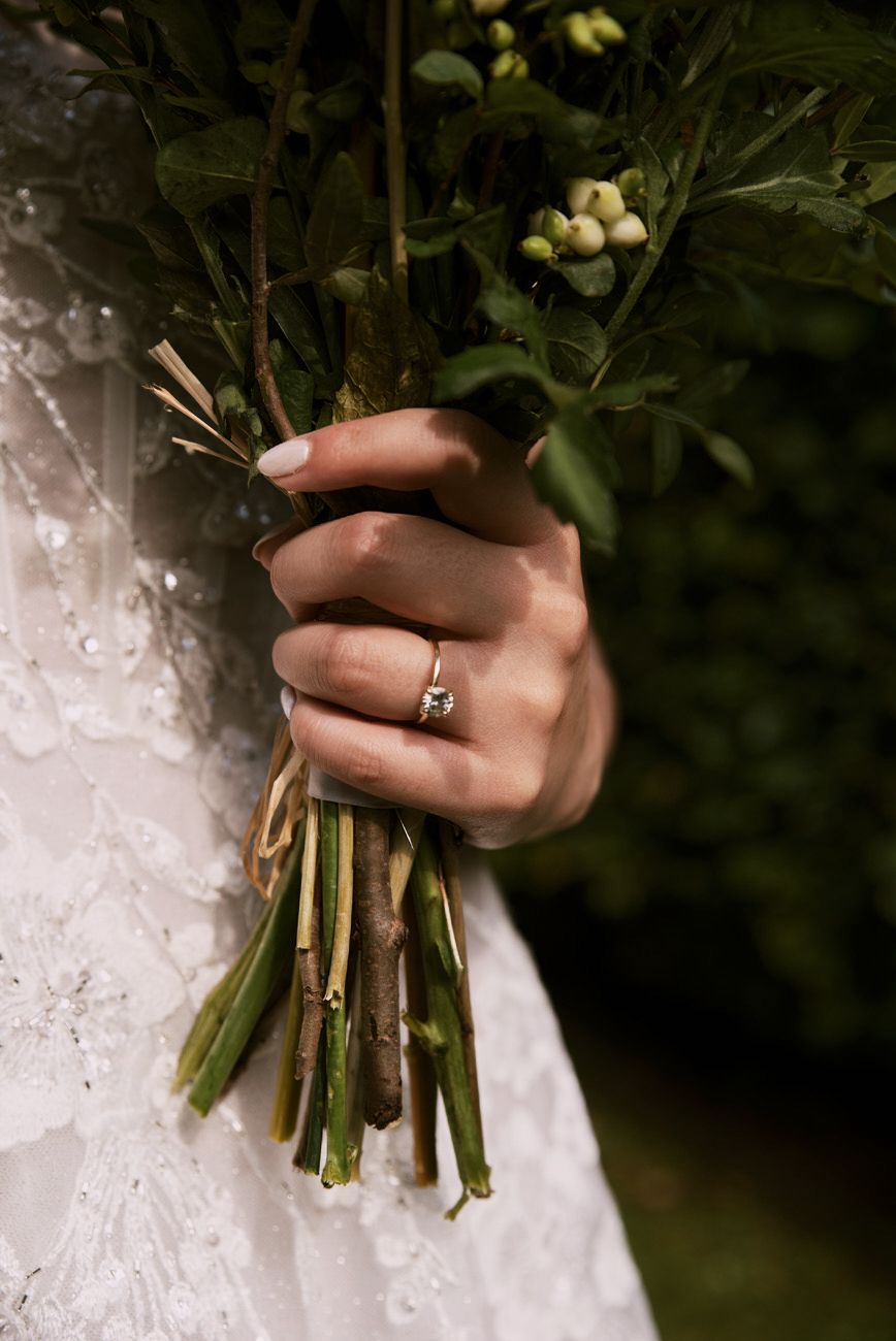 Bride holding a green botanical wedding bouquet with white berries, wearing a diamond engagement ring and lace wedding dress.