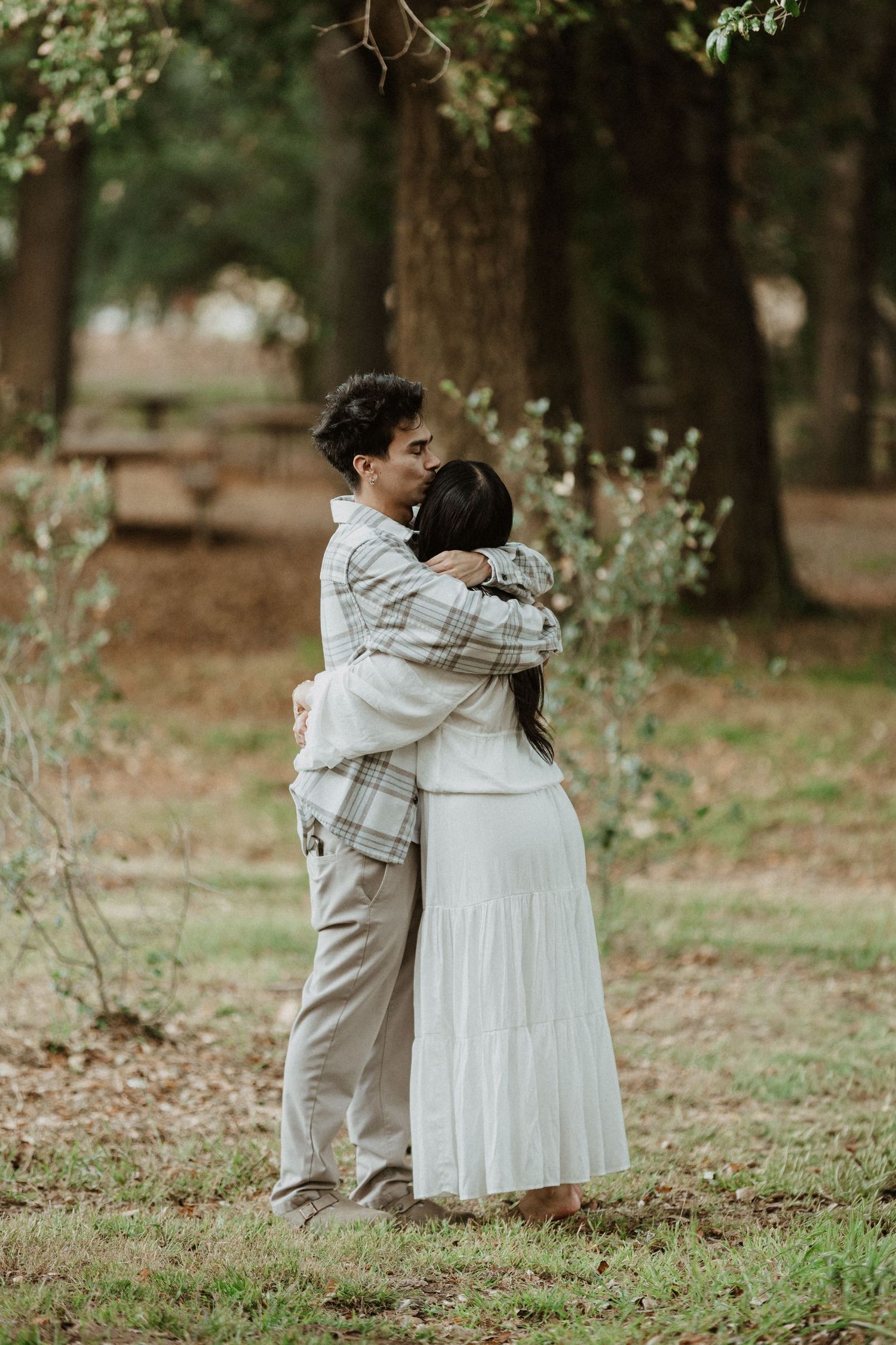 Couple embracing in a lush green forest, woman in white dress and man in plaid shirt sharing a romantic hug outdoors.