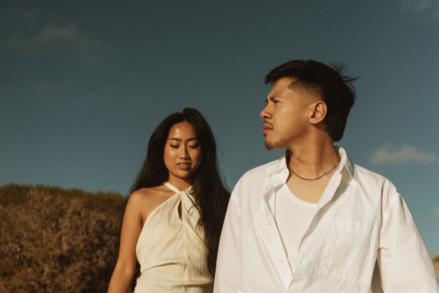 A young Asian couple stands outdoors under a blue sky, woman in beige halter dress, man in white shirt.