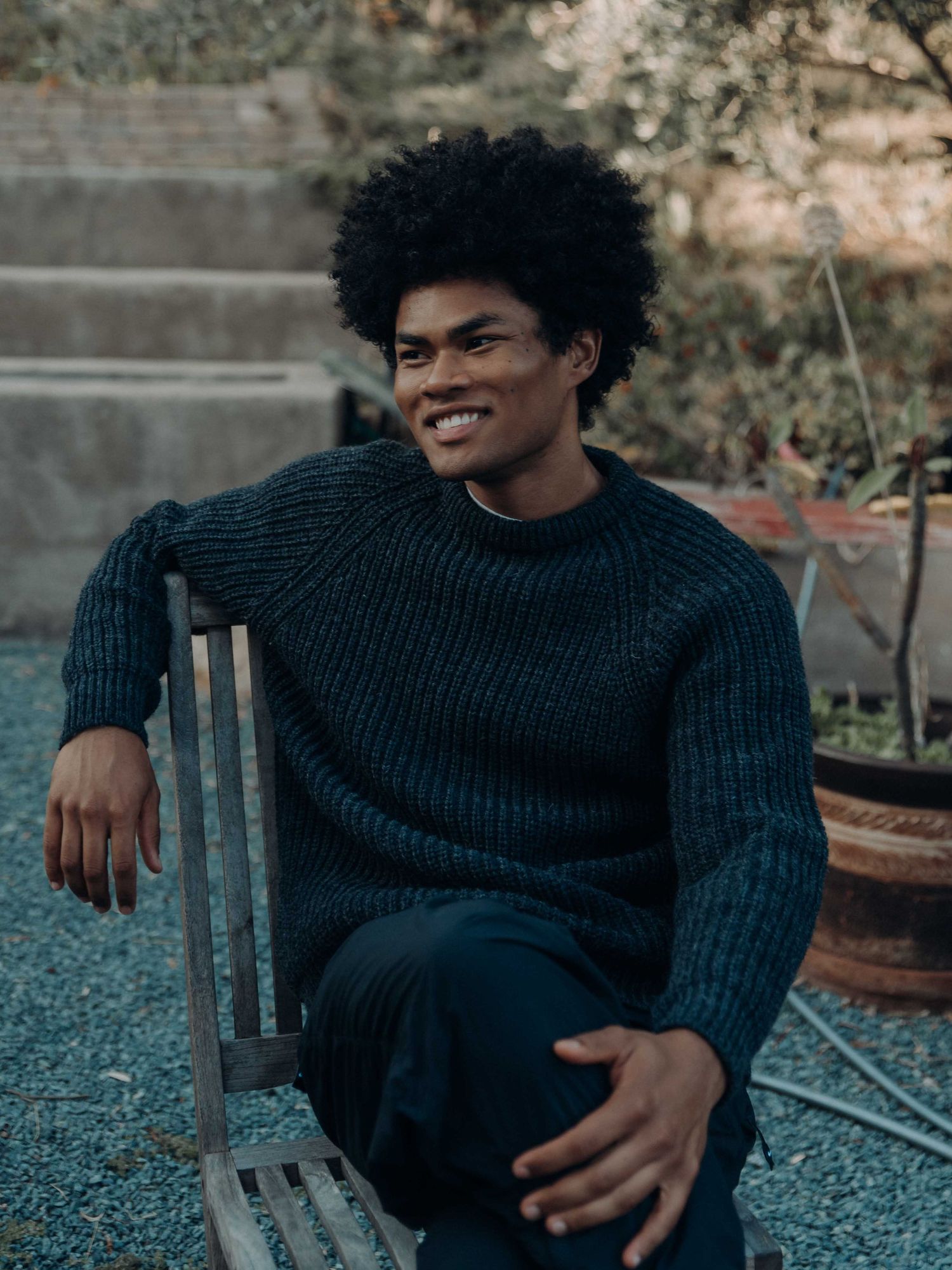 Young Black man with afro smiling, wearing dark knit sweater, seated on wooden chair in garden setting.