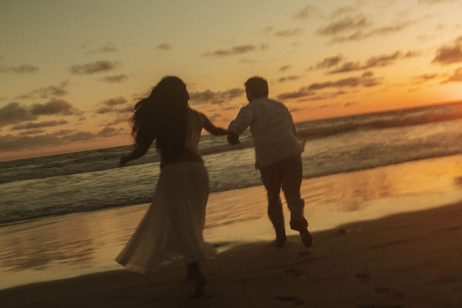 Couple holding hands running on a sandy beach at golden sunset with waves and colorful sky in the background.