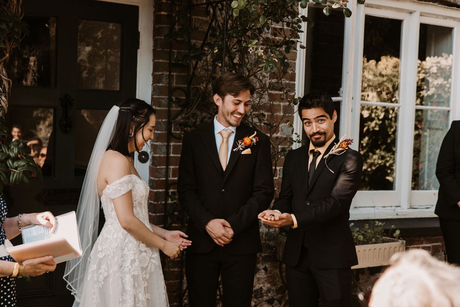 Groom laughs joyfully during outdoor wedding ceremony while officiant and guests look on smiling.