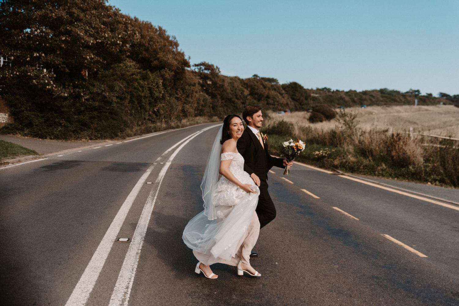 Bride and groom walking hand in hand across an open road, bride in white gown with veil.