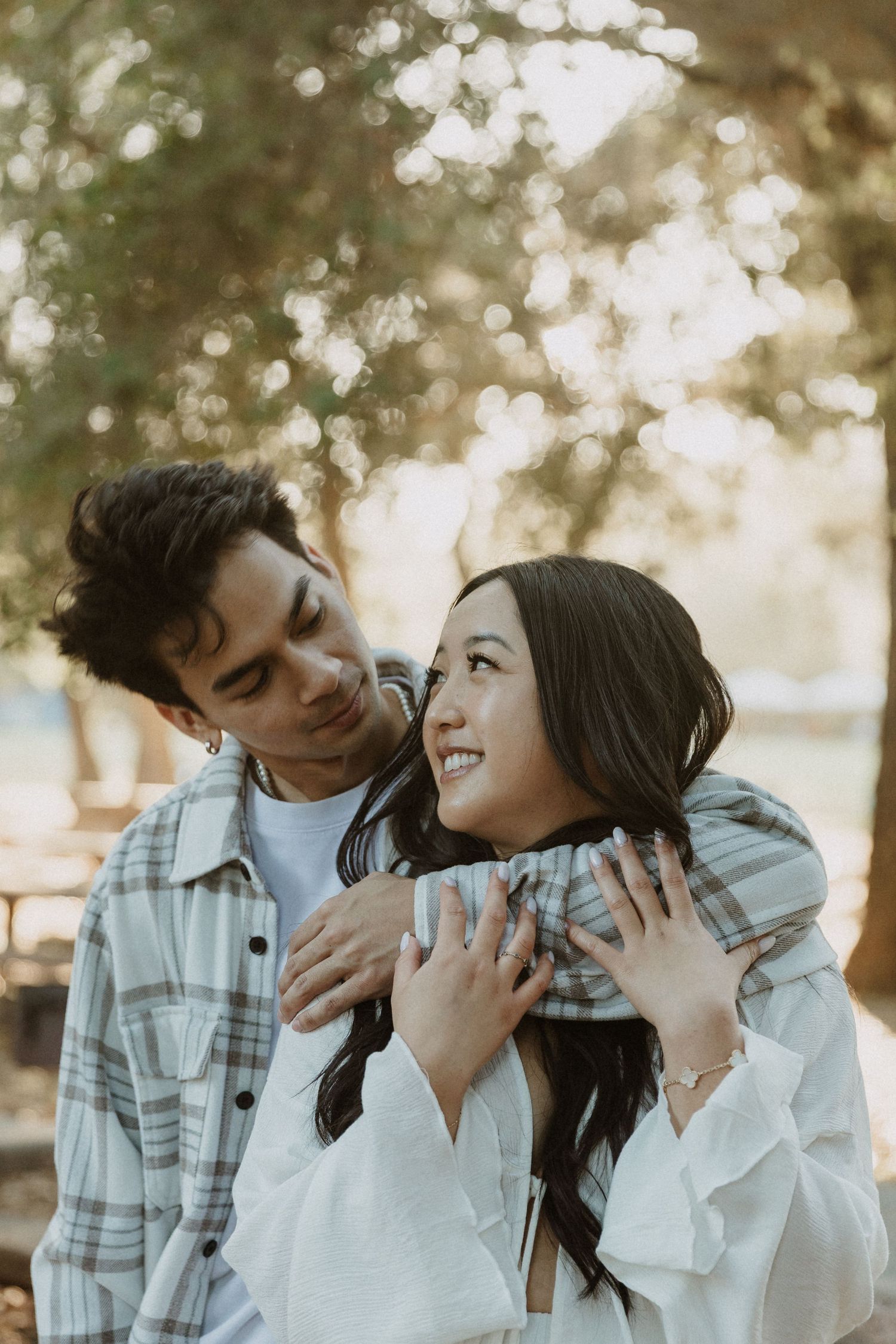 A young couple embraces outdoors, smiling at each other among blurred trees in warm natural light.