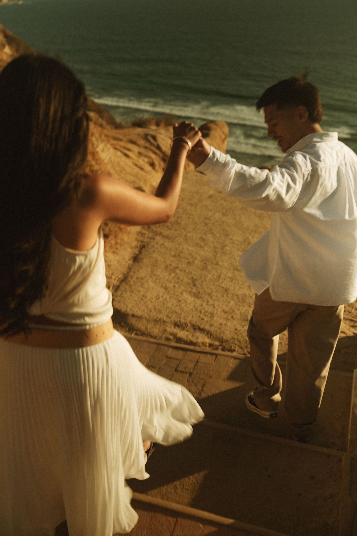 Couple dancing together on coastal cliffside steps at golden hour, woman in white dress, man in white shirt.
