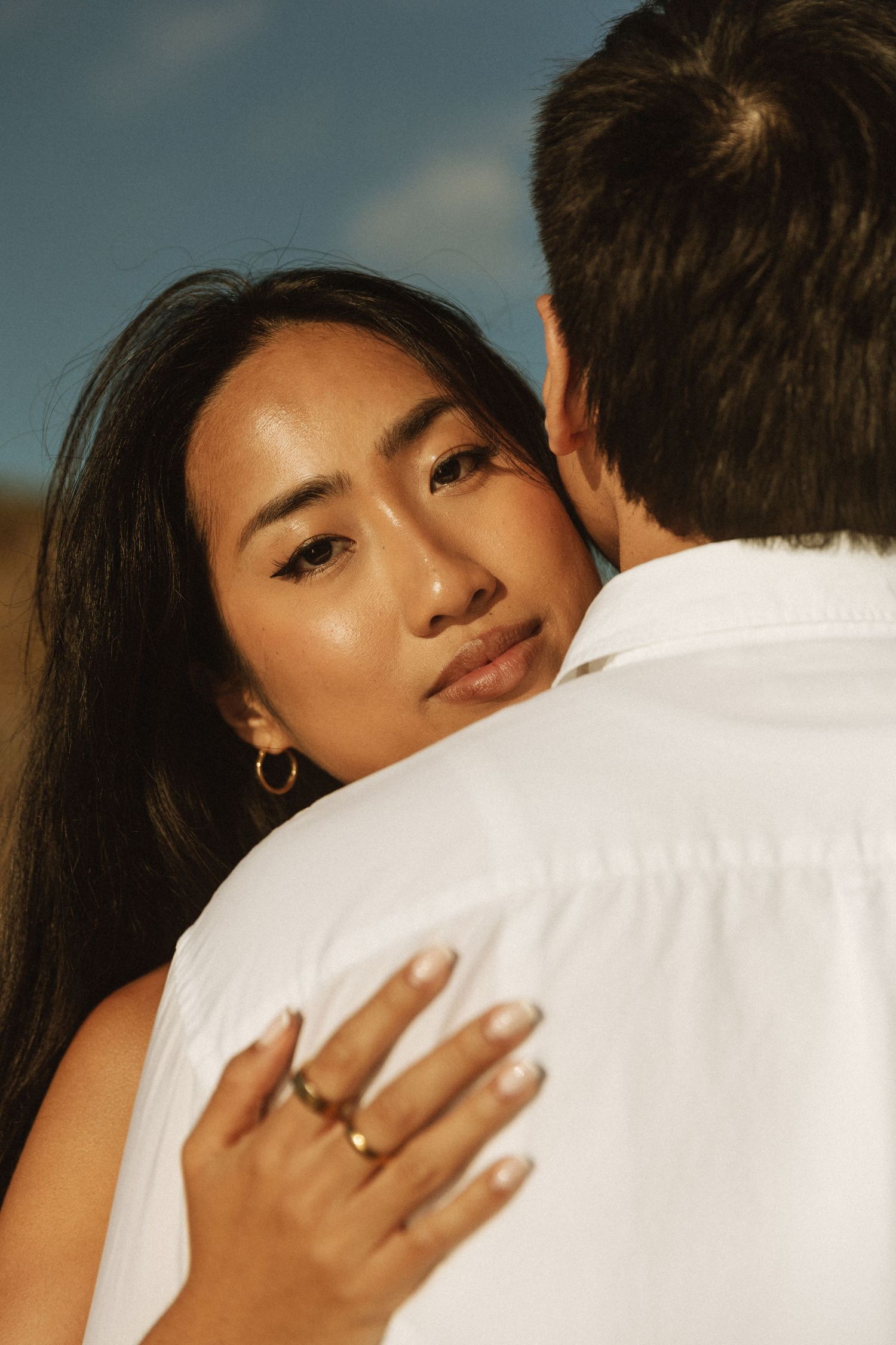 A woman with long dark hair embraces a man in a white shirt outdoors, wearing a gold ring and hoop earring.