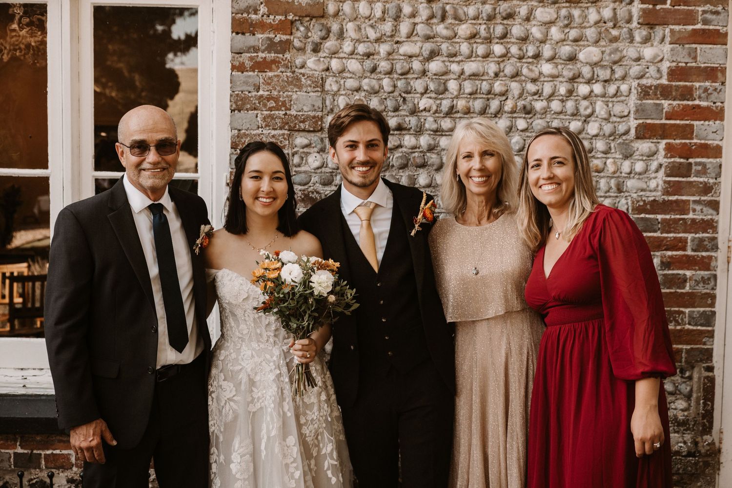 Wedding party of five posing outdoors against rustic brick wall, bride holding bouquet in white gown.