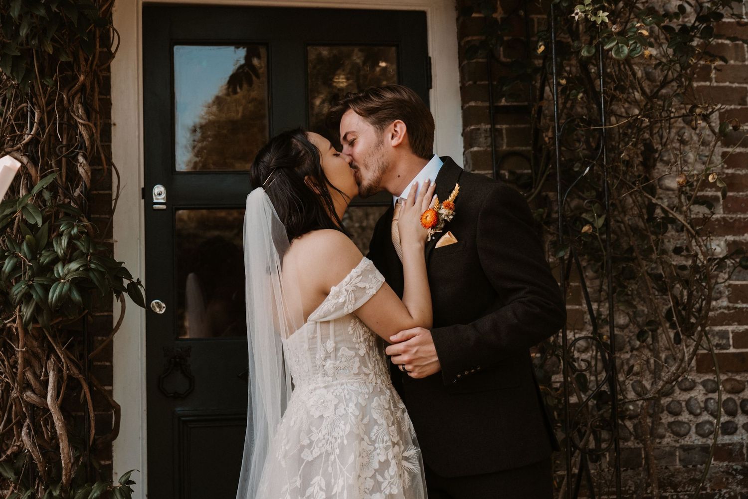 Newlywed couple shares first kiss in front of ivy-covered black door at brick wall venue.