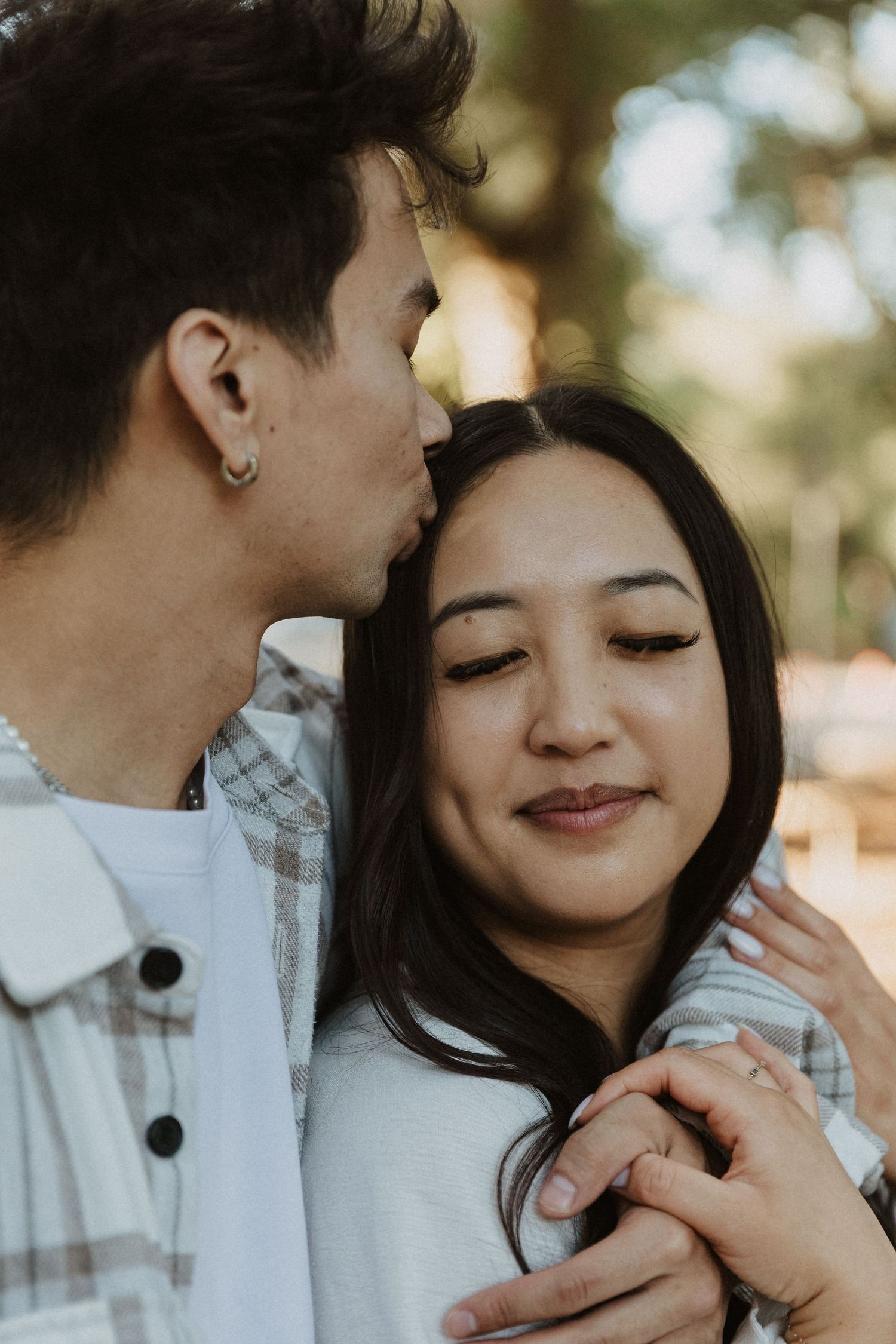 A young couple shares a tender moment outdoors, man kissing woman on temple while holding hands, both smiling softly.